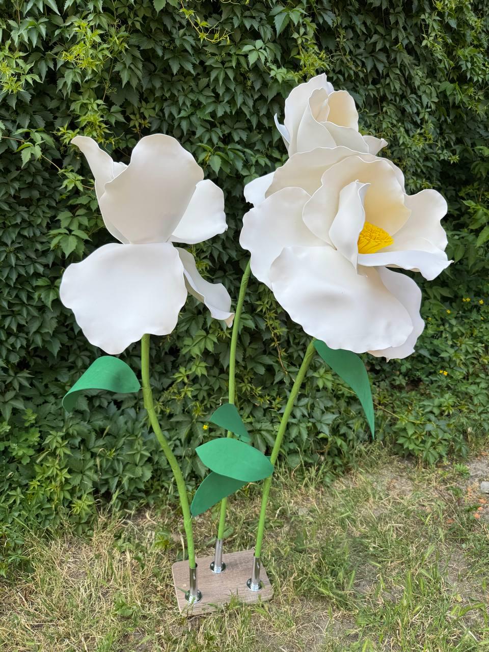 May include: Three large, oversized artificial white flowers with smooth, rounded petals and bright yellow centers. Each flower is attached to a thin green stem with green leaves, and all three stems are mounted into a rectangular wooden base.