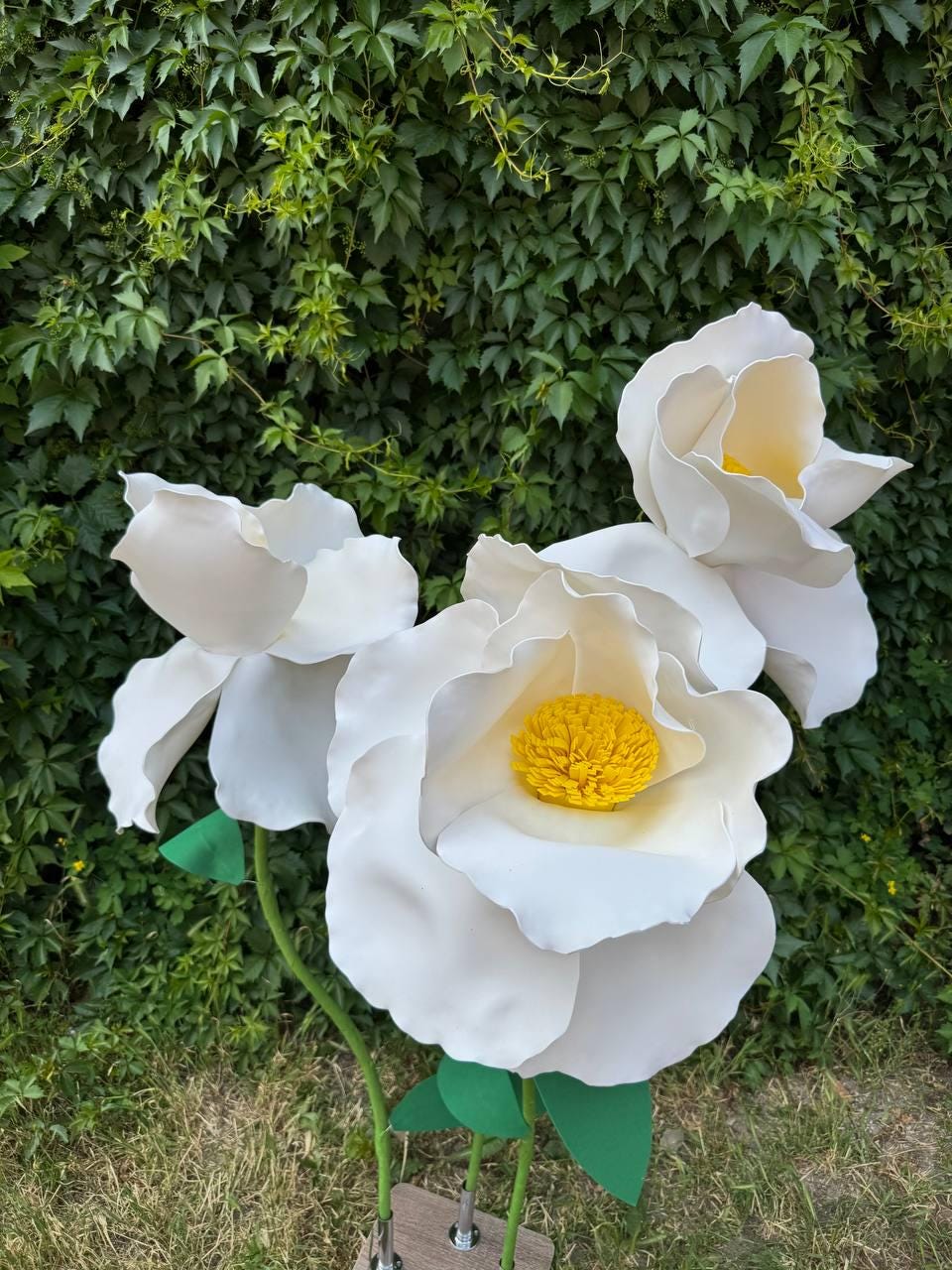 May include: Three large, oversized artificial flowers with white, rounded petals and bright yellow, textured centers. Each flower is attached to a thin green stem with green leaves, and the stems are mounted on a small, rectangular wooden base.