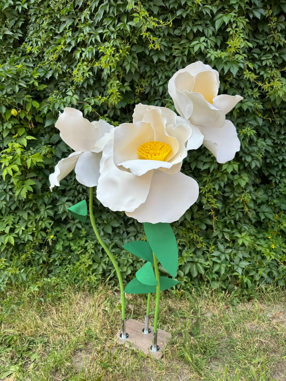 May include: Three large, oversized artificial flowers with white petals and bright yellow centers. Each flower is attached to a thin green stem with green leaves, and all three stems are mounted into a rectangular wooden base with metal supports.