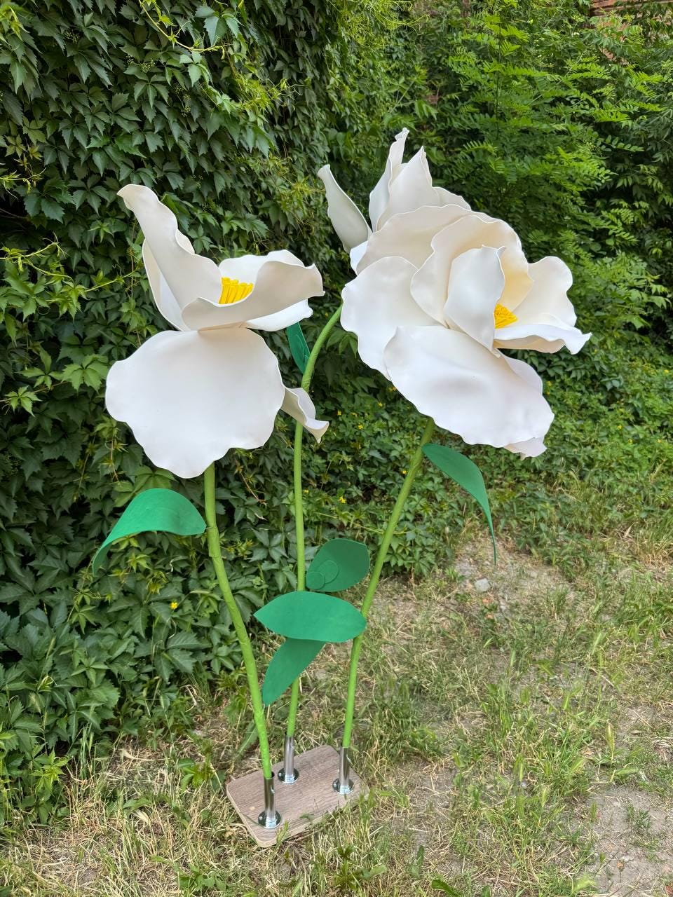 May include: Two large, oversized white artificial flowers with yellow centers, mounted on thin green stems with green leaves. The stems are attached to a rectangular wooden base, creating a freestanding decorative floral display.