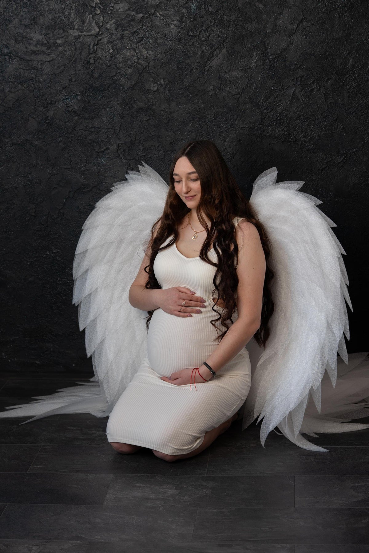 May include: A pregnant individual with long brown hair wearing a white ribbed sleeveless dress, kneeling while wearing large, white, layered feather-like wings that extend outward on both sides against a dark, textured background.