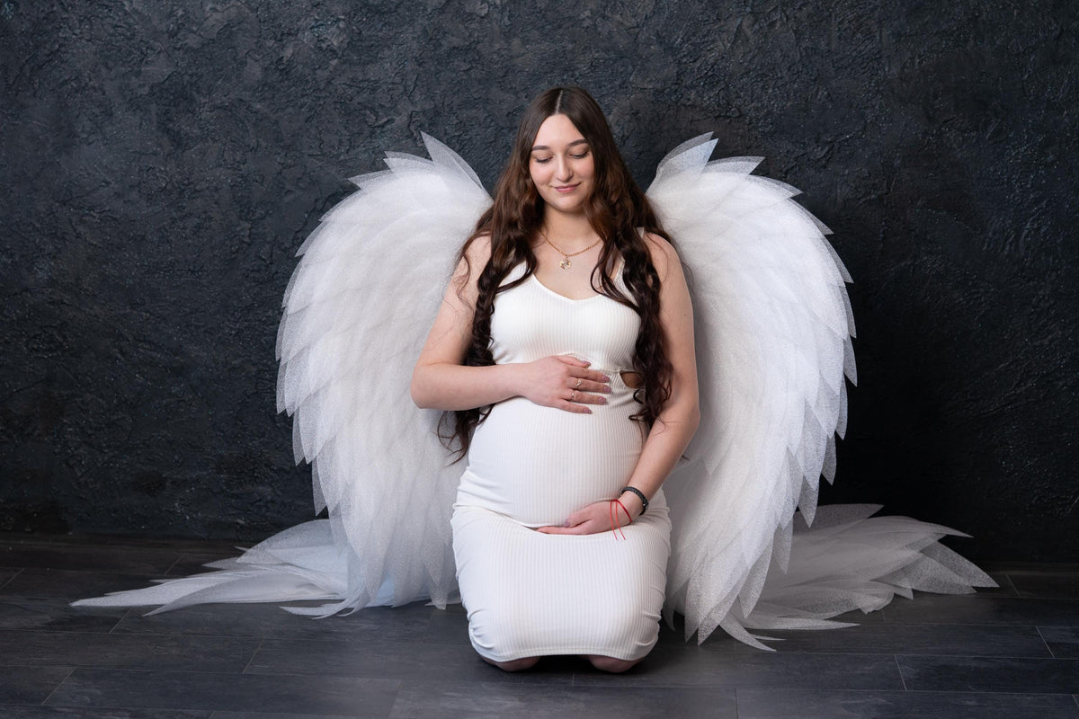 May include: A pregnant individual wearing a white ribbed sleeveless dress, kneeling with large, white, layered feather-like wings attached to the back. The wings extend outward and downward, creating a dramatic, expansive silhouette against a dark background.