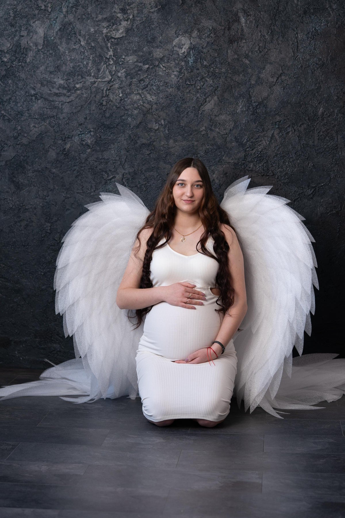 May include: A white ribbed sleeveless dress worn by a pregnant individual, featuring large, layered, translucent white angel wings attached to the back. The garment has a scoop neckline and a fitted silhouette that extends to the knees.