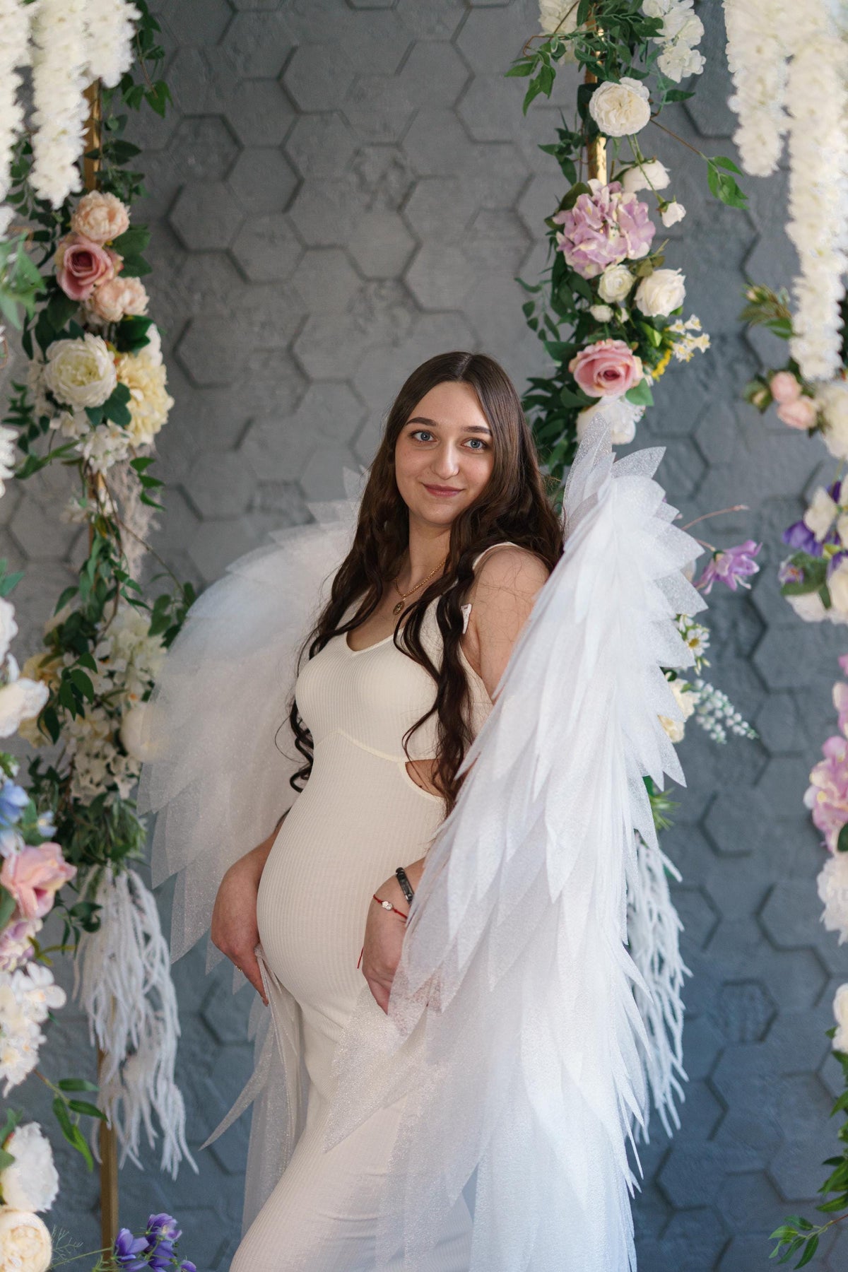May include: A cream-colored ribbed maternity dress with side cutouts, paired with large, layered white tulle angel wings. The dress features a V-neckline and a form-fitting silhouette that highlights the baby bump, creating a soft and ethereal look.