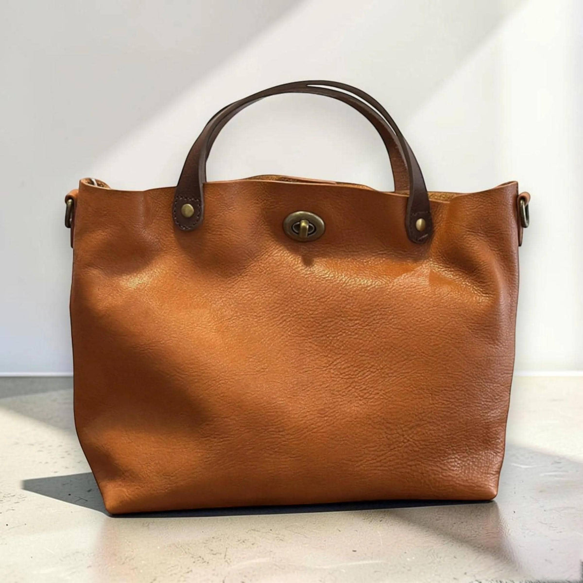 May include: A tan leather handbag featuring a smooth, textured surface, two dark brown leather handles attached with brass rivets, and a central brass turn-lock closure. The bag has a wide, open-top design with side rings for a shoulder strap.