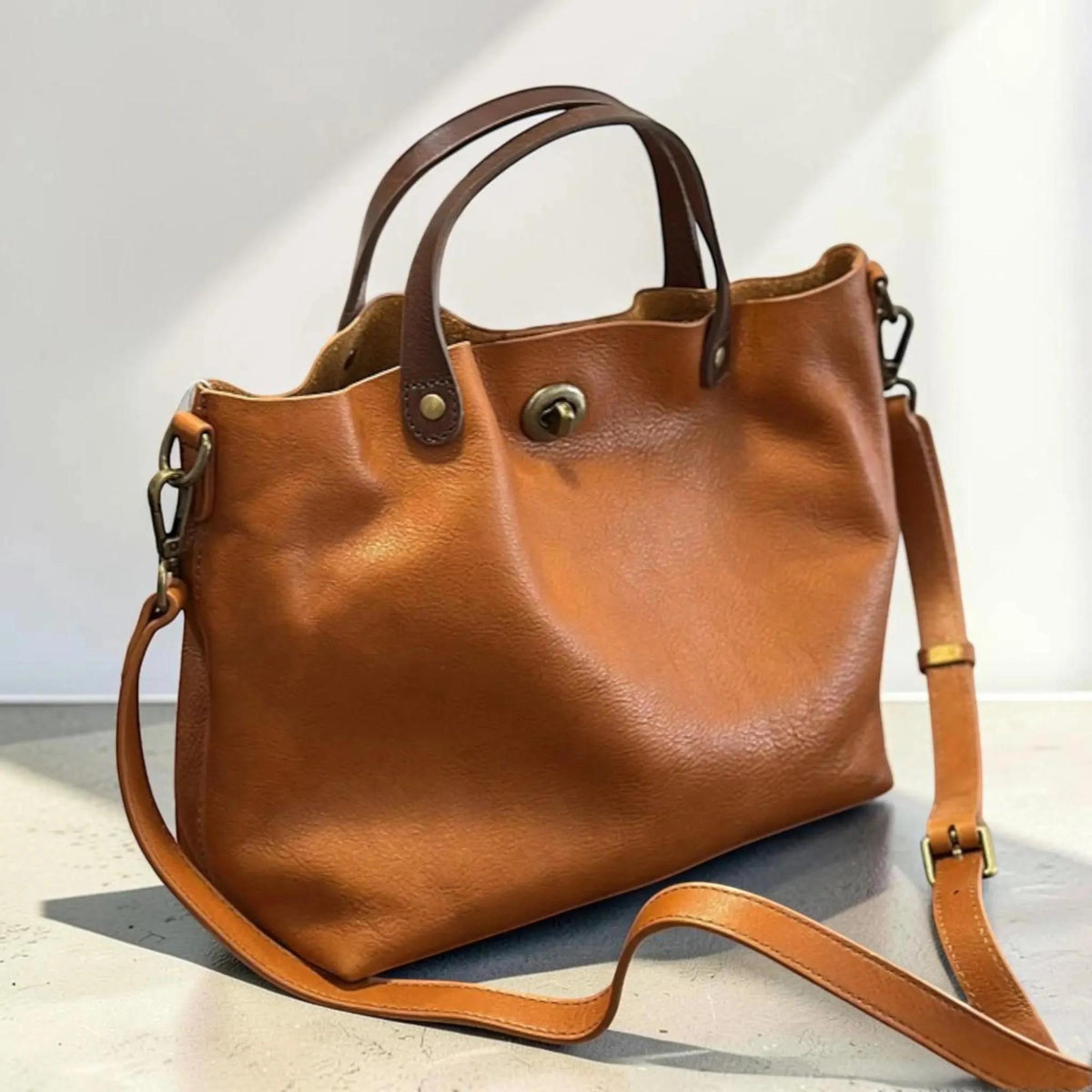 May include: A tan leather handbag featuring two dark brown top handles and a detachable shoulder strap. The bag has a central brass turn-lock closure, antique brass hardware, and a textured surface. The design includes side clips for the strap.