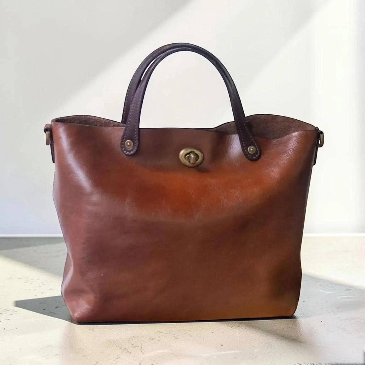 May include: A medium-sized brown leather tote bag featuring two dark brown handles attached with brass rivets. The bag has a central brass turn-lock closure on the front panel. The leather has a smooth, polished finish with visible natural grain.