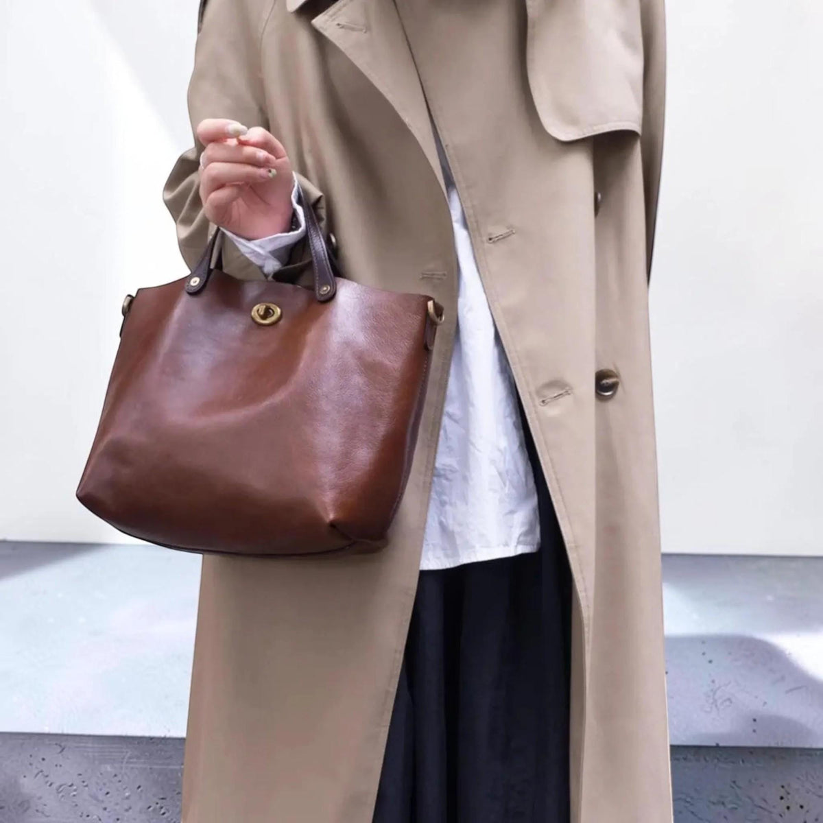 May include: A dark brown leather tote bag featuring a gold-toned metal turn-lock closure at the top center. The bag has two dark brown leather handles and is worn with a beige trench coat, a white collared shirt, and a black skirt.