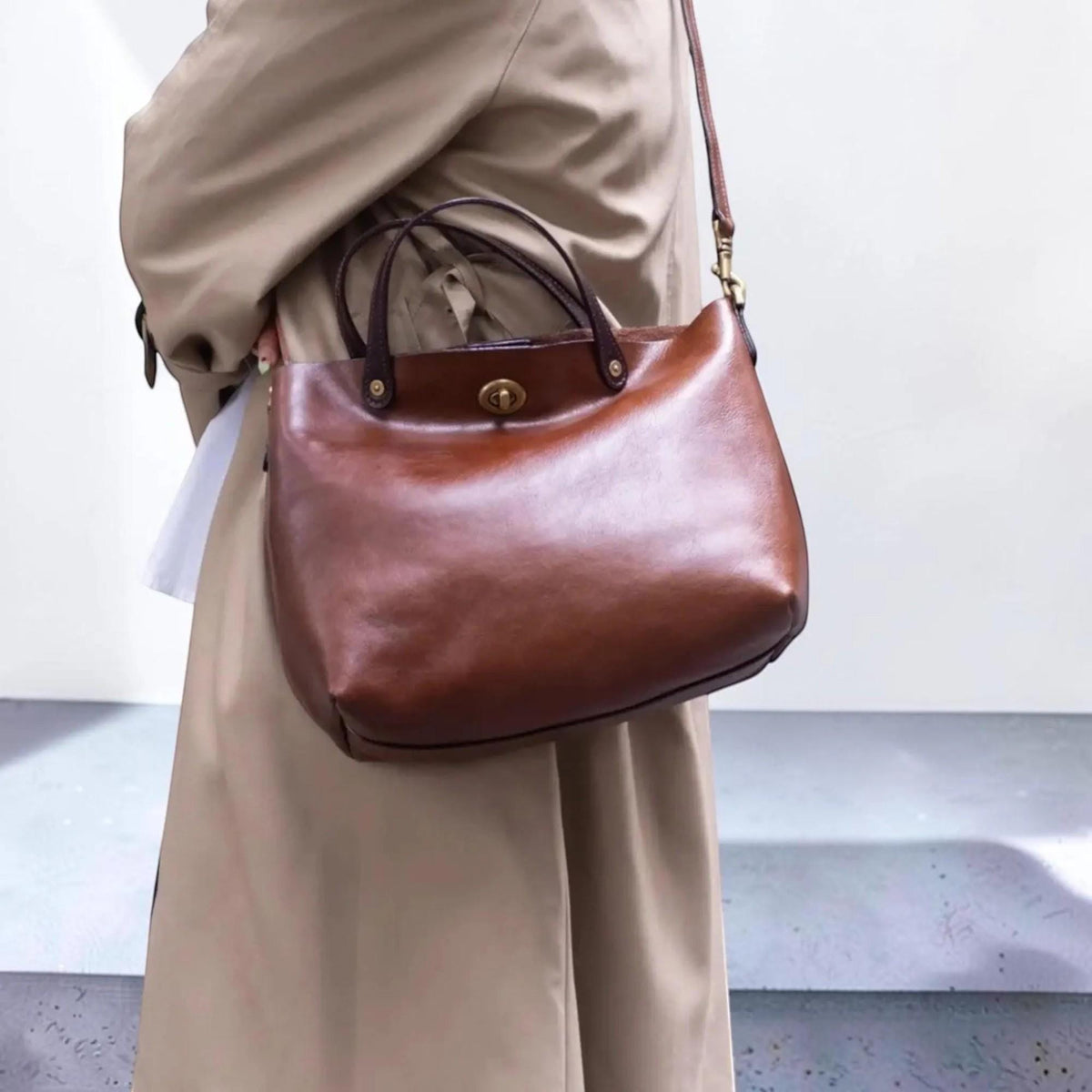 May include: A smooth brown leather handbag featuring two dark brown top handles and a brass-toned metal turn-lock closure. The bag includes a thin, adjustable leather shoulder strap attached with brass-toned hardware, providing a versatile carry option.