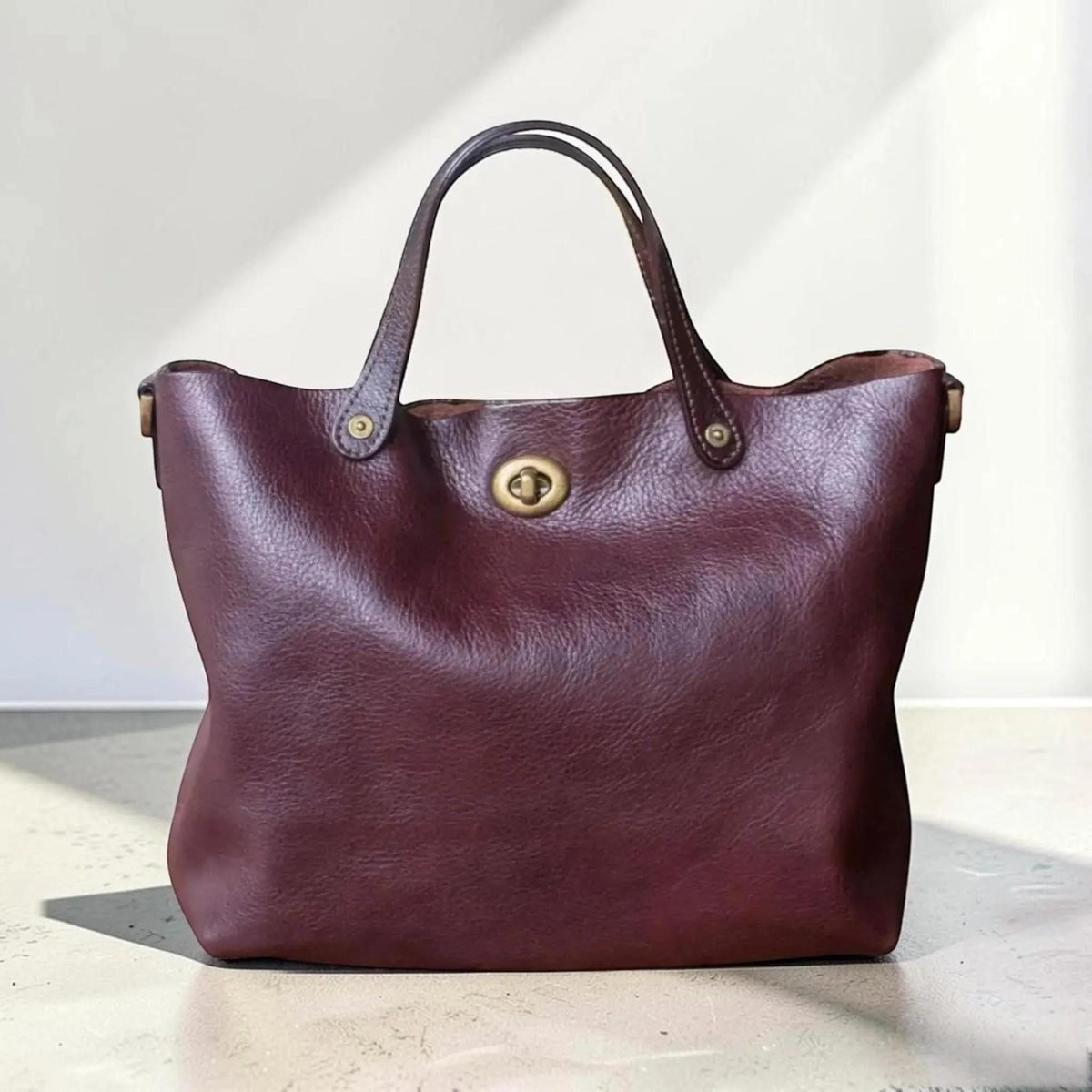May include: A deep burgundy leather tote bag featuring a textured surface, two sturdy top handles, and a central brass-toned metal turn-lock closure. The bag has a wide, open-top design with visible stitching details at the handle attachments.