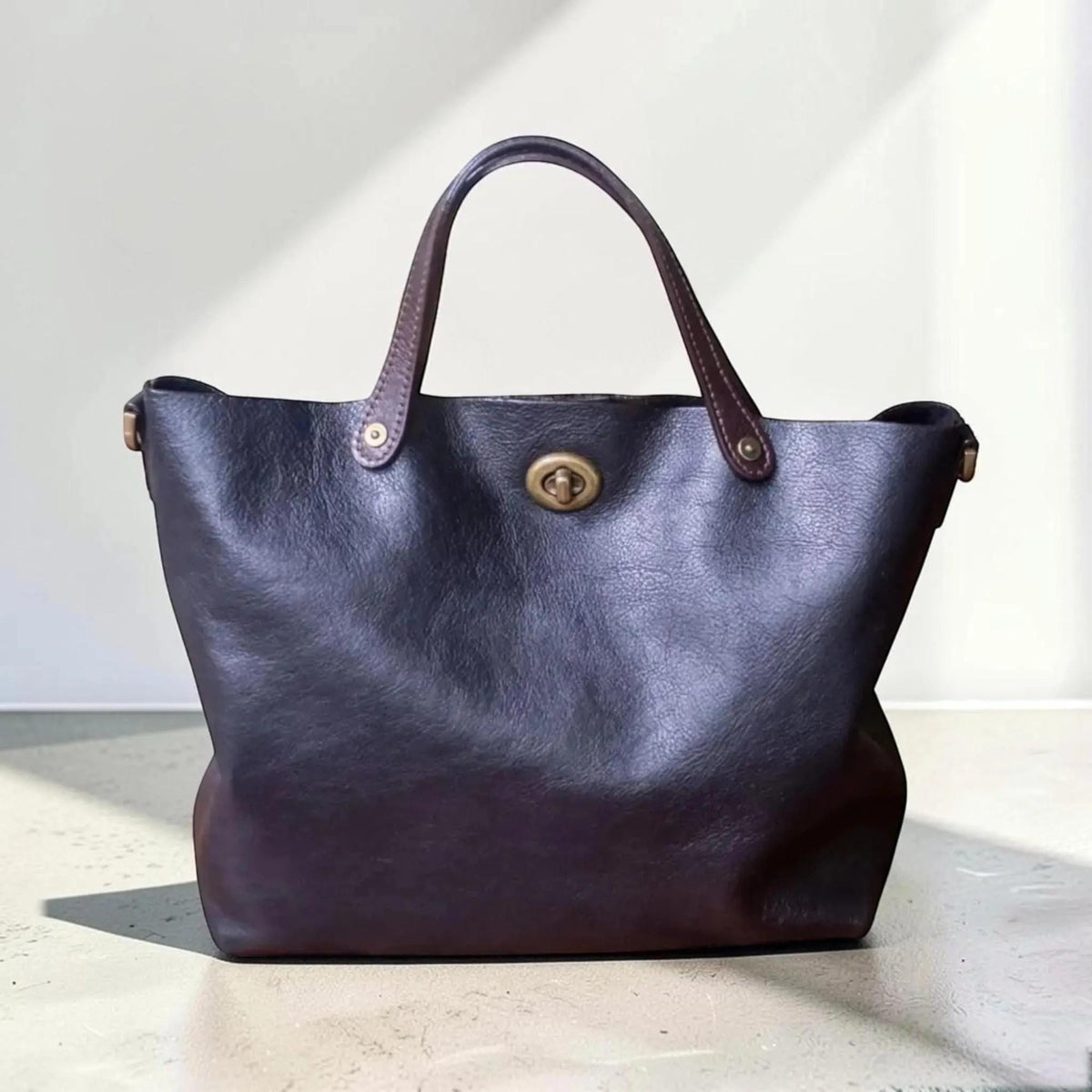 May include: A dark brown leather tote bag featuring two sturdy handles attached with brass rivets and a central brass turn-lock closure. The bag has a soft, unstructured silhouette with a visible grain texture throughout the dark brown material.