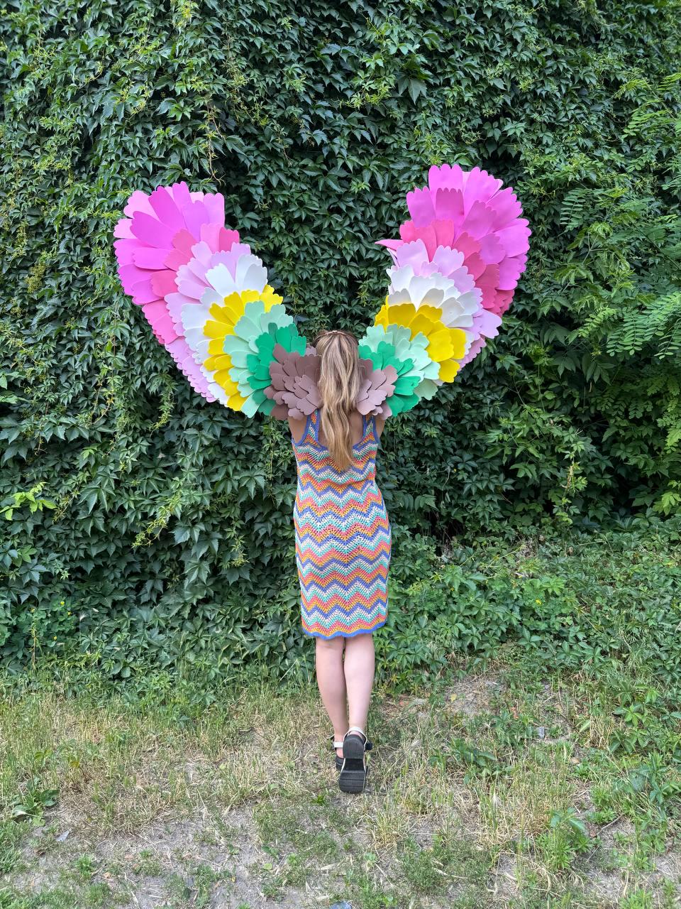 May include: Large decorative wings constructed from layered paper feathers in a vibrant rainbow gradient of pink, yellow, green, and brown. The wings are positioned behind a sleeveless dress featuring a multi-colored chevron pattern in orange, blue, and white.