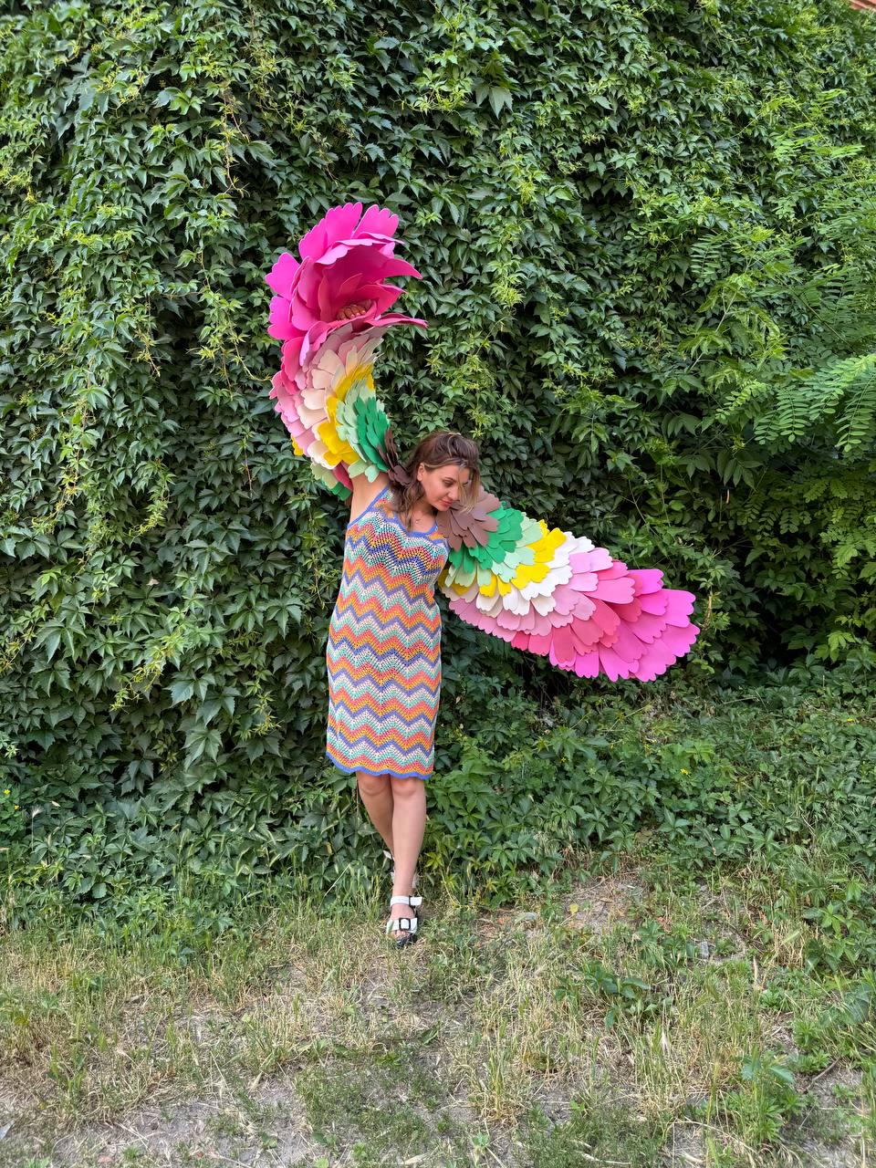 May include: A sleeveless chevron-patterned knit dress in orange, blue, and white, paired with large, wearable bird wings featuring layered, scalloped feathers in shades of pink, yellow, green, and brown, worn with white strappy sandals.