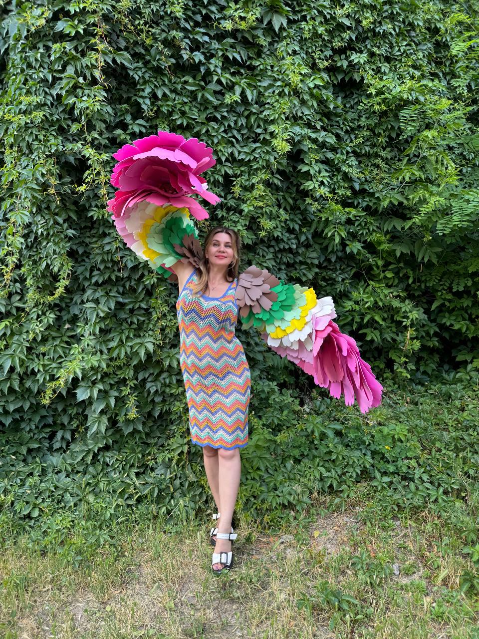 May include: A sleeveless knee-length dress featuring a multi-colored chevron pattern in orange, blue, yellow, and green. Attached to the shoulders are large, wing-like structures made of layered, scalloped fabric petals in shades of pink, green, and brown.