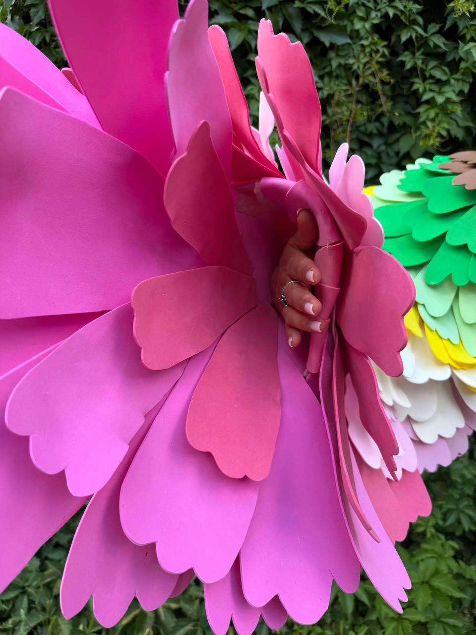 May include: A large, oversized decorative flower crafted from multiple layers of pink foam petals with scalloped edges. The structure features a central opening, and the petals are arranged in a dense, overlapping pattern to create a voluminous, three-dimensional floral shape.