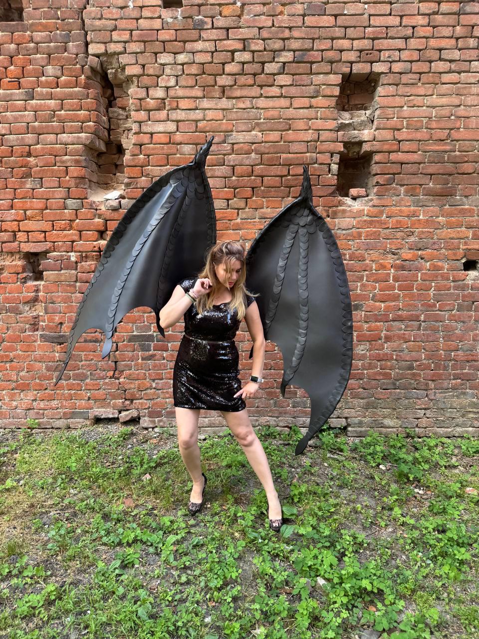 May include: A black sequined mini dress paired with large, articulated black dragon wings featuring a textured, scale-like pattern. The wings are tall and pointed, extending upwards and outwards, while the dress is sleeveless with a fitted silhouette.