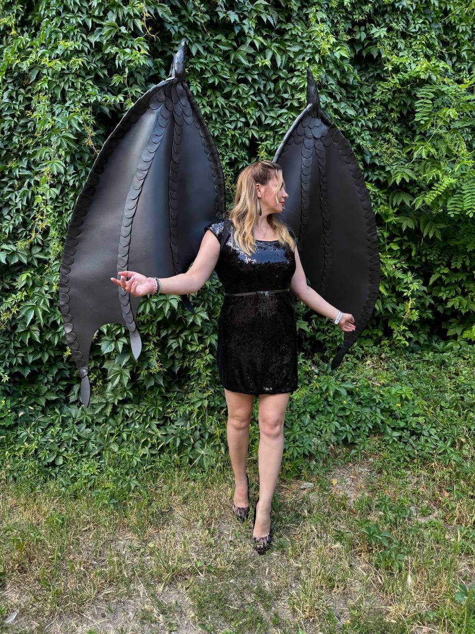 May include: A pair of large, black, articulated dragon wings featuring a layered, scale-like texture along the edges, worn with a black sequined mini dress that has a scoop neckline and a thin belt at the waist, paired with black high-heeled shoes.