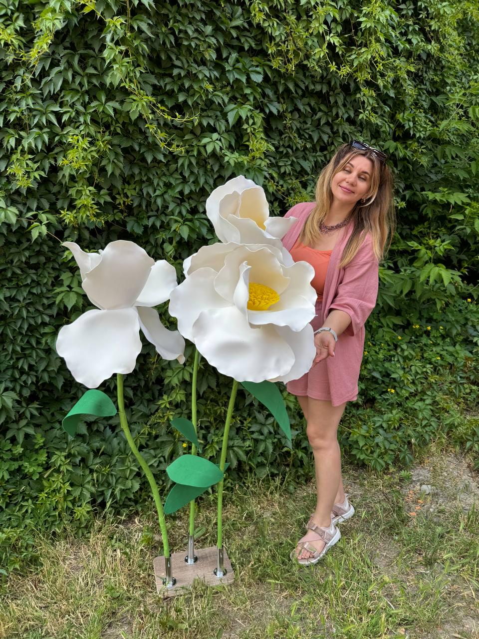 May include: Two oversized artificial white flowers with yellow centers, mounted on thin green stems with green leaves. The flowers are attached to a rectangular base with metallic connectors, standing upright as a decorative floor display piece.