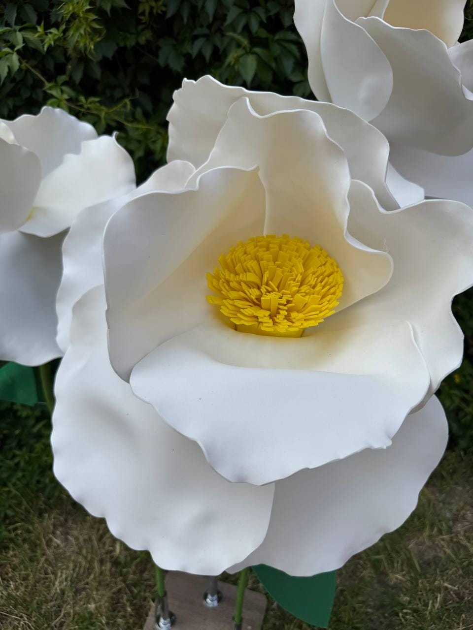 May include: Large decorative artificial flowers featuring oversized white petals with smooth, wavy edges and a bright yellow, textured center. The flowers are mounted on thin green stems, showcasing a realistic floral design with multiple layers.