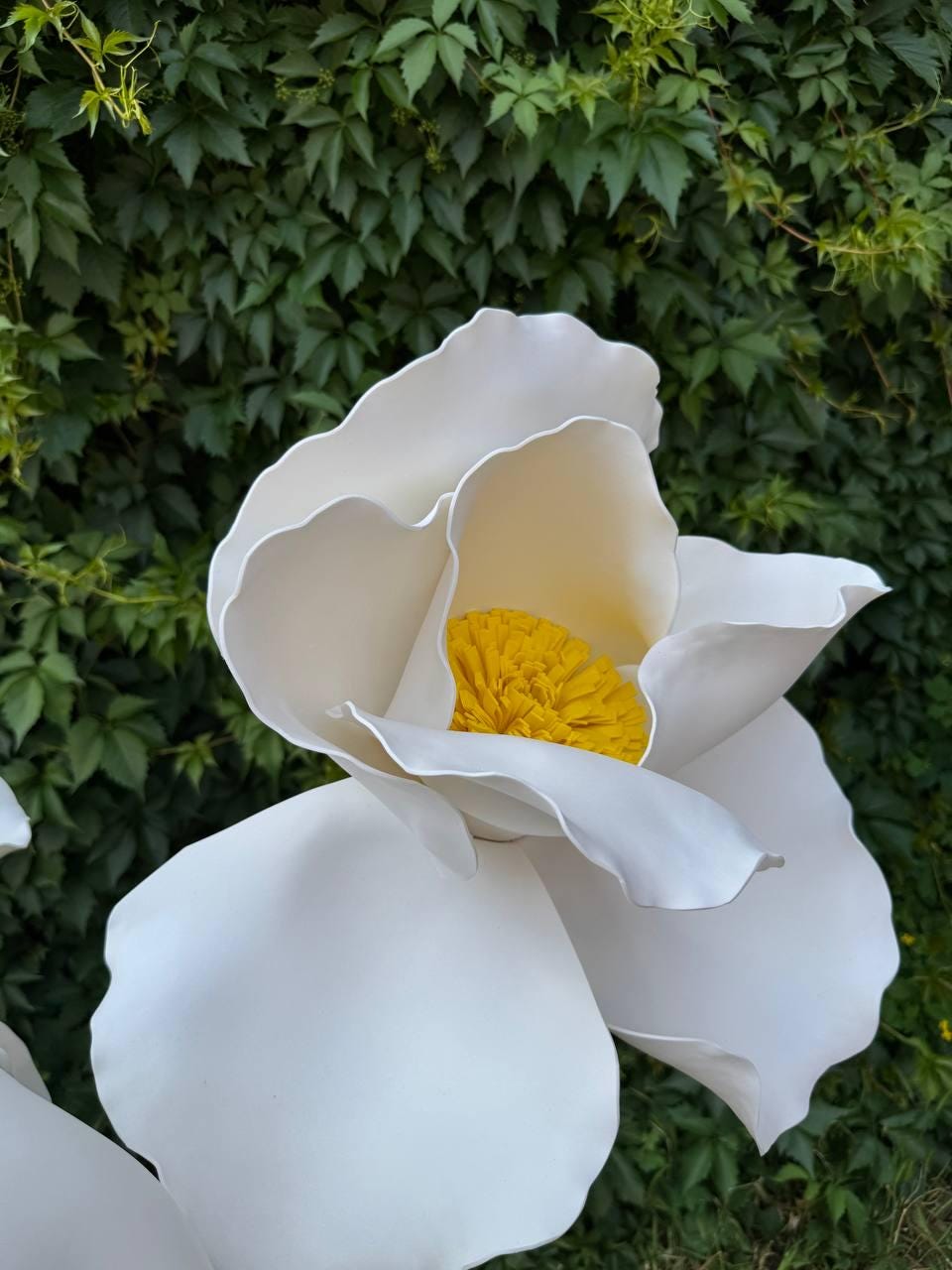 May include: A large, decorative artificial flower featuring smooth, matte white petals with wavy, organic edges. The center of the flower is composed of a dense, textured cluster of bright yellow stamens, creating a high-contrast focal point.