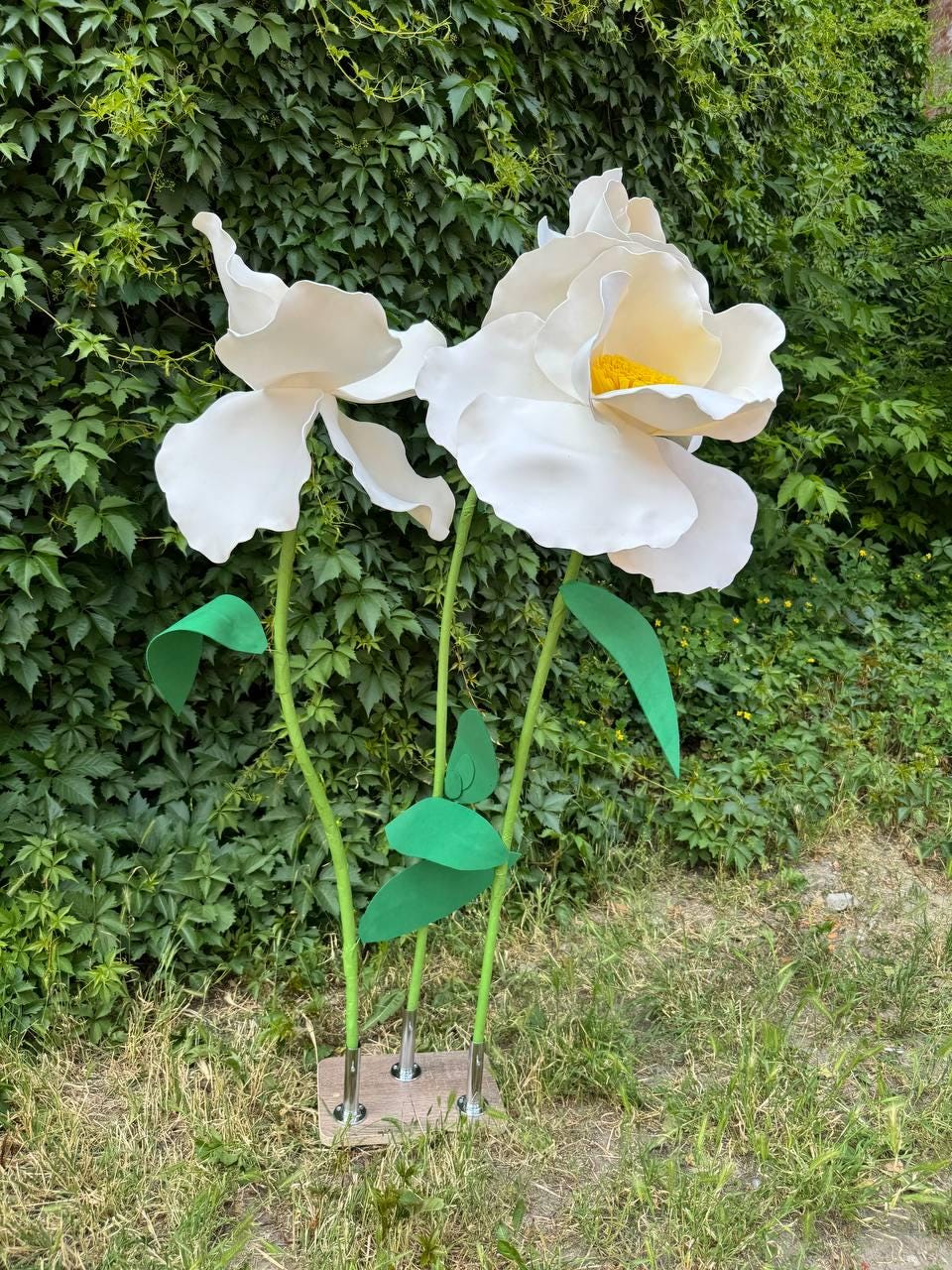 May include: Two large, oversized artificial white flowers with yellow centers, mounted on thin green stems with green leaves. The flowers are attached to a rectangular base with metal connectors, standing upright as a decorative floral display.