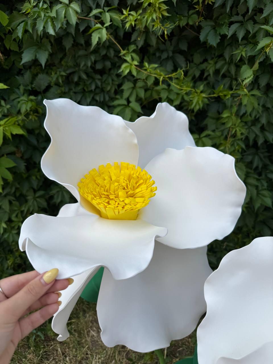 May include: A large, oversized decorative flower featuring smooth, white, rounded petals with wavy edges and a bright yellow, textured, fringed center. The flower is constructed from a firm material and displays a bold, high-contrast appearance.