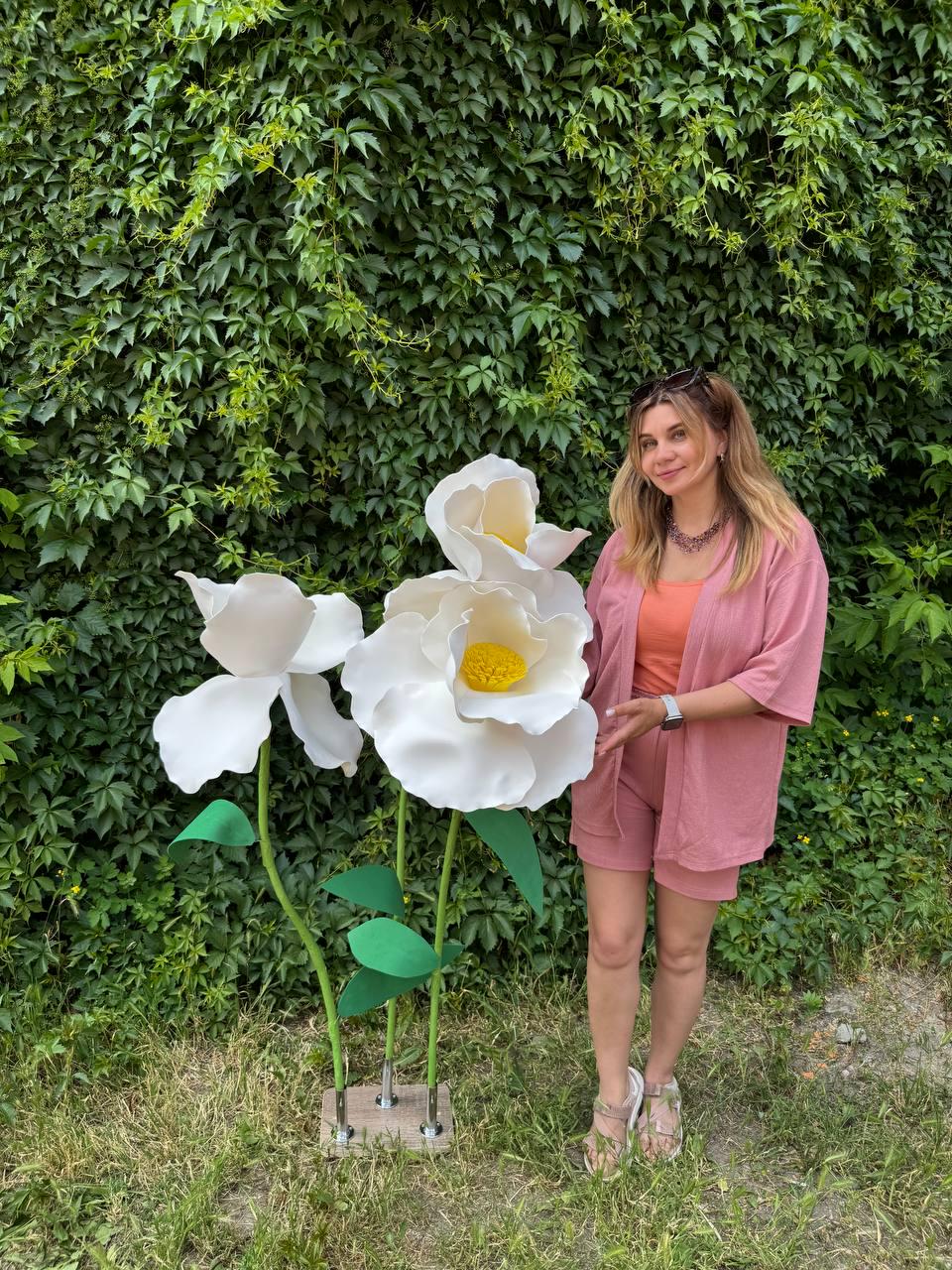 May include: Two oversized white artificial flowers with yellow centers, green stems, and green leaves, mounted on a square base. The flowers feature large, layered petals and are displayed alongside a person wearing a pink cardigan and shorts.