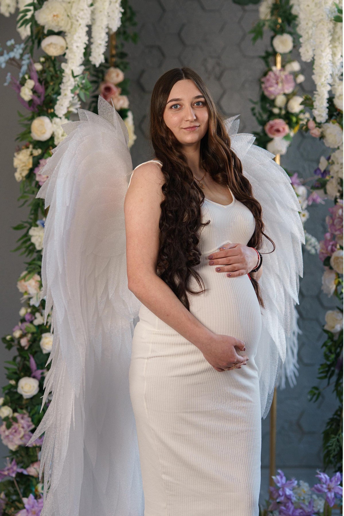 May include: A white ribbed sleeveless dress worn with large, translucent white angel wings featuring layered, feather-like structures. The dress is form-fitting, reaching mid-calf, and features a scoop neckline. The wings are expansive and textured.