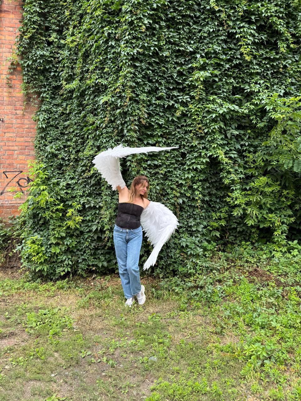 May include: Large white feathered wings attached to a black strapless corset top and light blue denim jeans. The wings are expansive and textured with layered white feathers, extending outward from the back of the black bodice and blue pants.