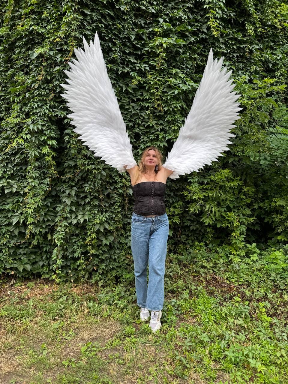 May include: Large white feathered angel wings attached to a black strapless corset top, blue denim jeans, and white sneakers. The wings feature layered, pointed white feathers extending upwards and outwards, creating a wide, expansive span.