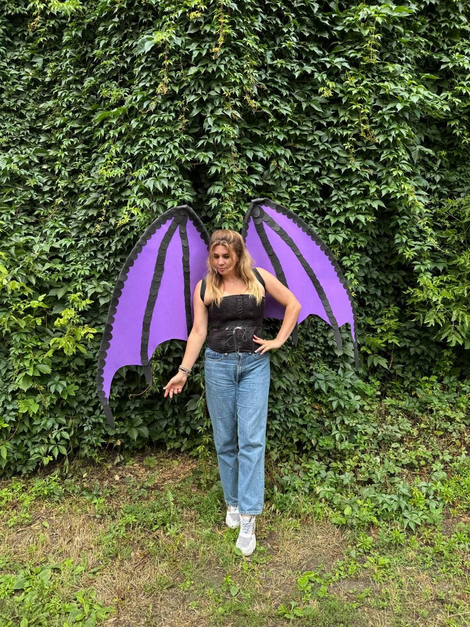 May include: A pair of large, purple dragon-style wings with black scalloped edges, worn over a black corset top and blue denim jeans. The wings feature a structured frame with distinct segments, extending outward from the back of the garment.