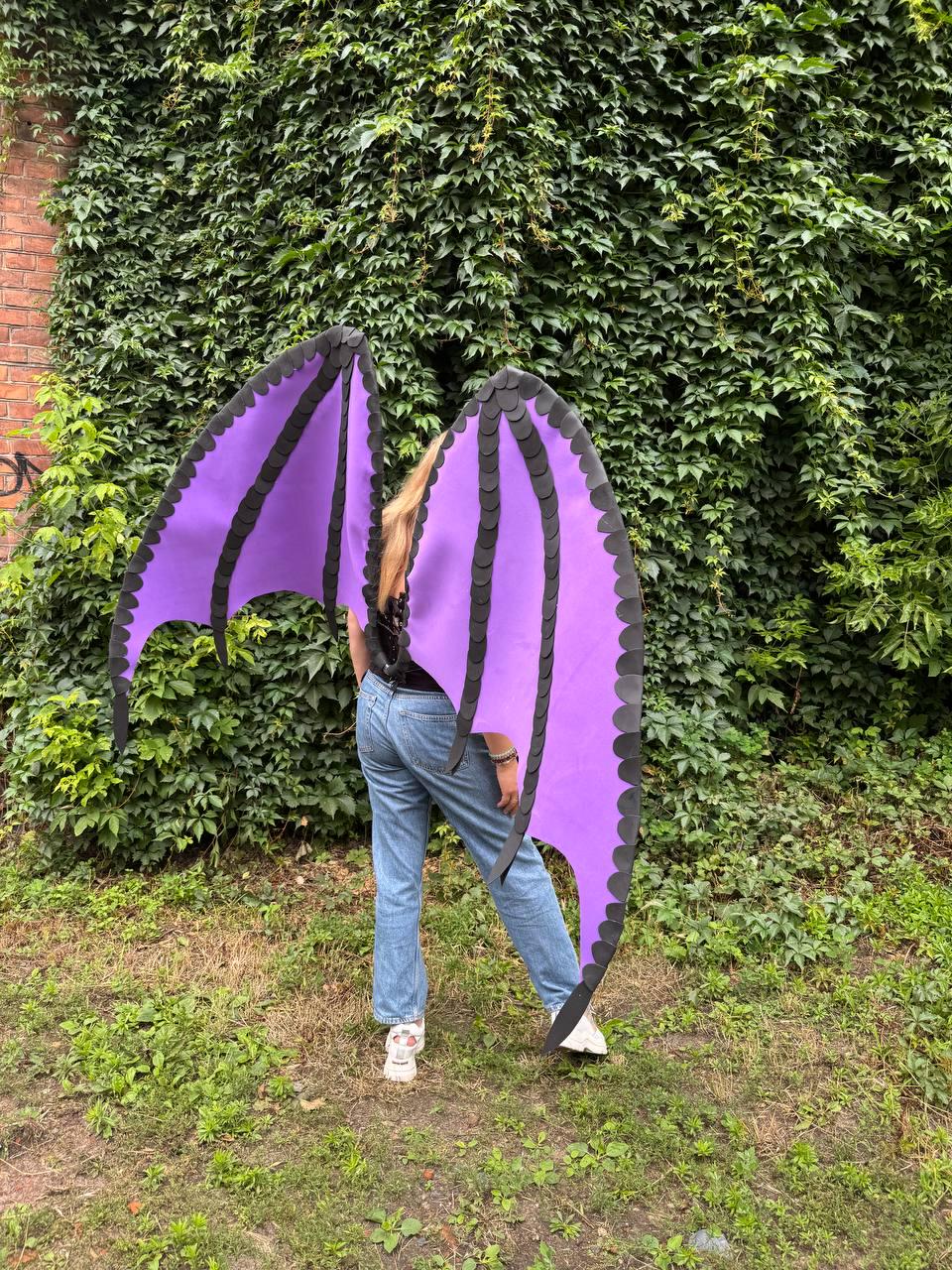 May include: Large, articulated dragon wings featuring a vibrant purple fabric membrane framed by black, scalloped-edge structural supports. The wings are designed to be worn on the back, showcasing a pointed, bat-like silhouette with detailed layering.
