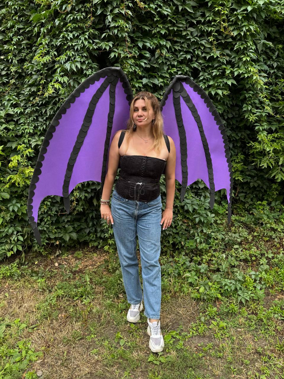May include: Large, pointed dragon-style wings featuring a vibrant purple membrane with black structural ribs and edges. The wings are worn over a black strapless corset top with front lacing and blue denim jeans, paired with white athletic sneakers.