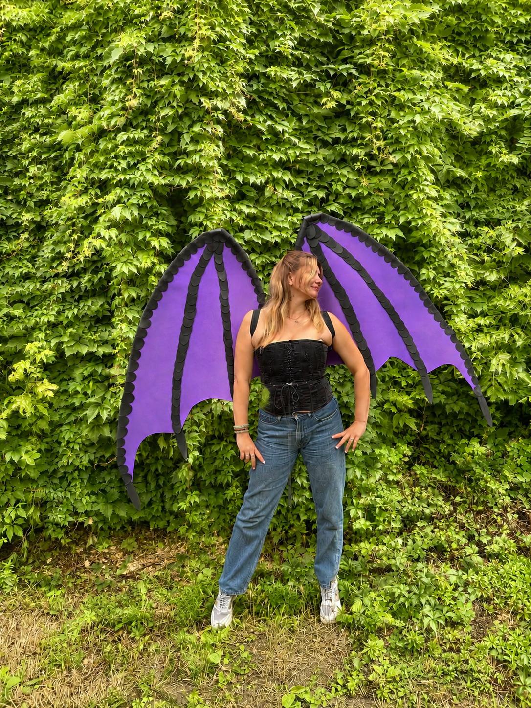 May include: Large, vibrant purple dragon wings with black structural detailing and scalloped edges, worn with a black lace-up corset top, blue denim jeans, and white sneakers. The wings feature a distinct bat-like shape with segmented panels.