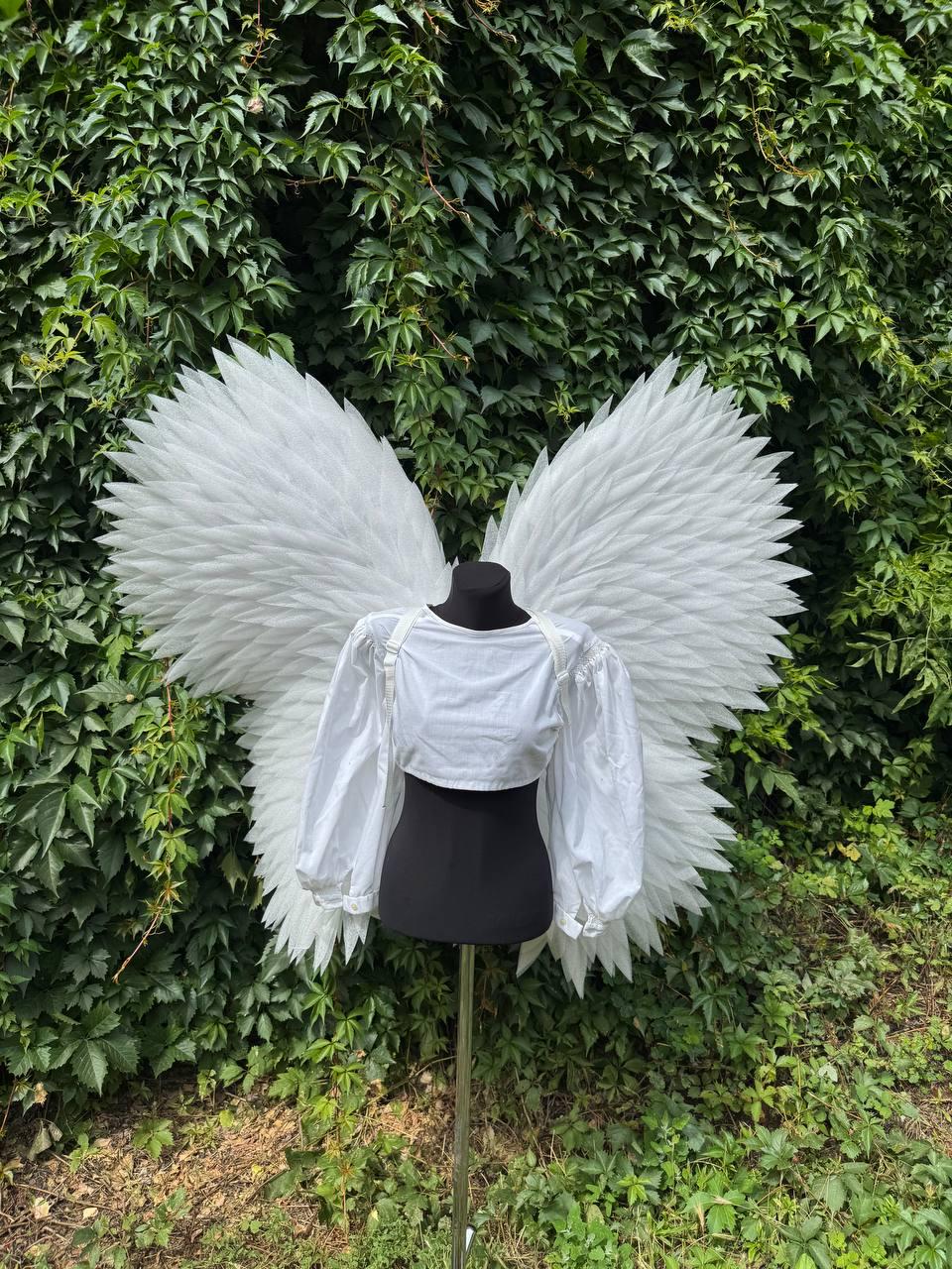 May include: Large white feathered angel wings attached to a black mannequin torso. The mannequin wears a white cropped long-sleeve shirt with puffed sleeves. The wings feature layered, pointed white feathers extending outward in a wide span.