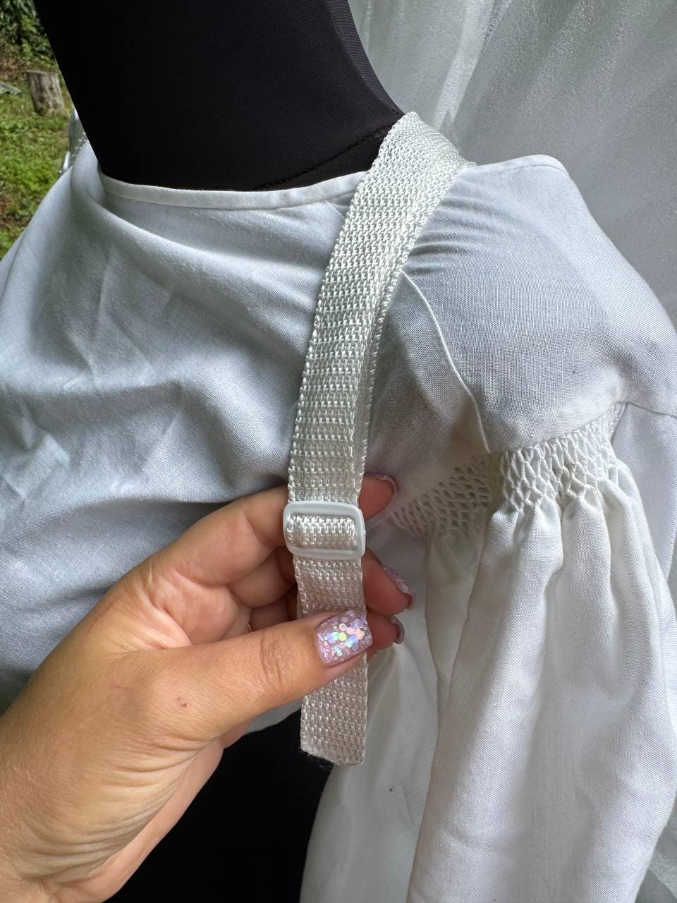 May include: A white blouse featuring gathered fabric details at the shoulder and a white adjustable woven strap with a plastic slider buckle. The garment is displayed on a mannequin, highlighting the texture of the fabric and the functional strap.