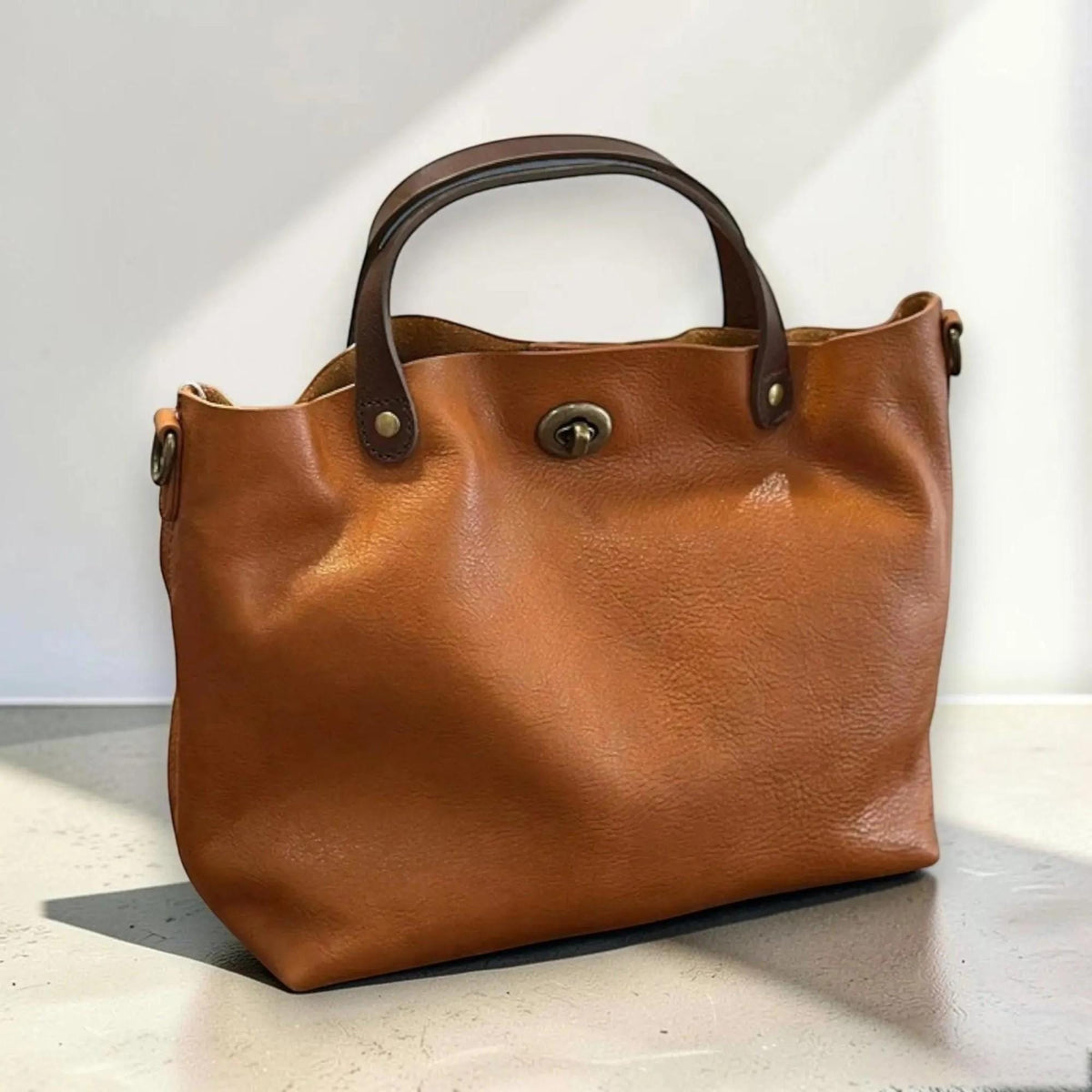 May include: A tan leather handbag featuring two dark brown top handles, a central antique brass turn-lock closure, and side metal rings for an optional strap. The bag has a soft, unstructured silhouette with visible grain texture throughout.