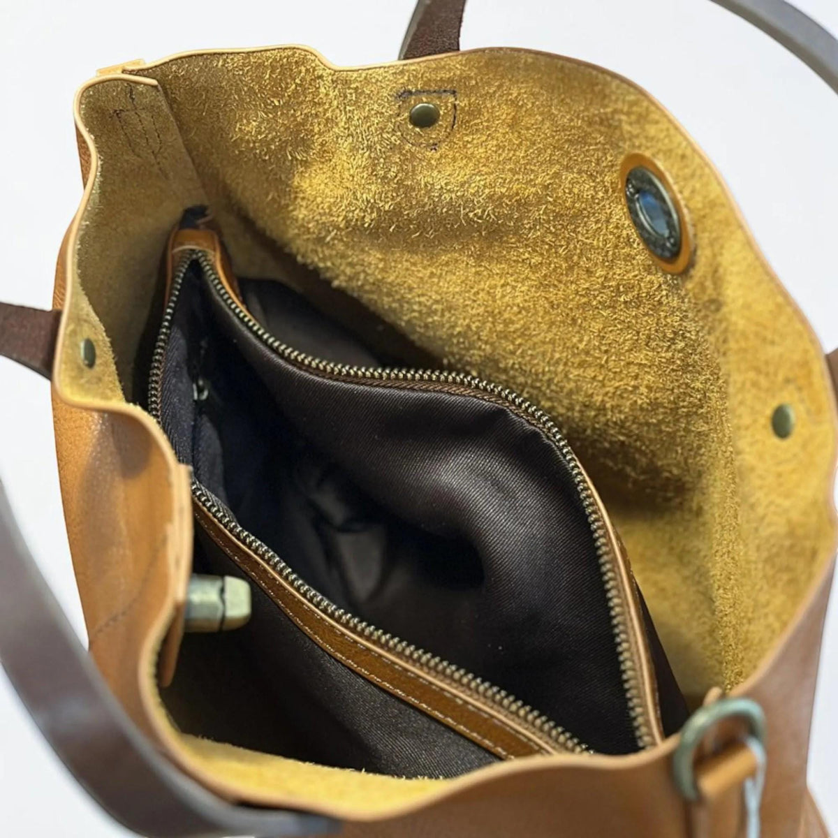 May include: A tan leather tote bag with a raw, unlined interior texture. The bag features dark brown leather handles, a central zippered compartment lined with dark brown fabric, and a metallic snap button closure at the top opening.