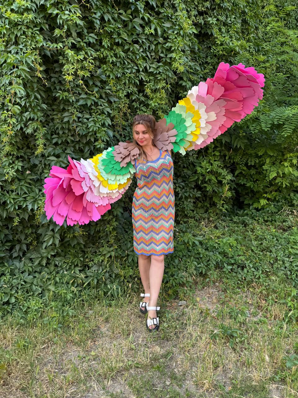 May include: A sleeveless, knee-length dress featuring a multi-colored chevron pattern in orange, blue, and yellow. Attached to the back are large, expansive wings constructed from layered, scalloped fabric petals in a vibrant rainbow gradient.