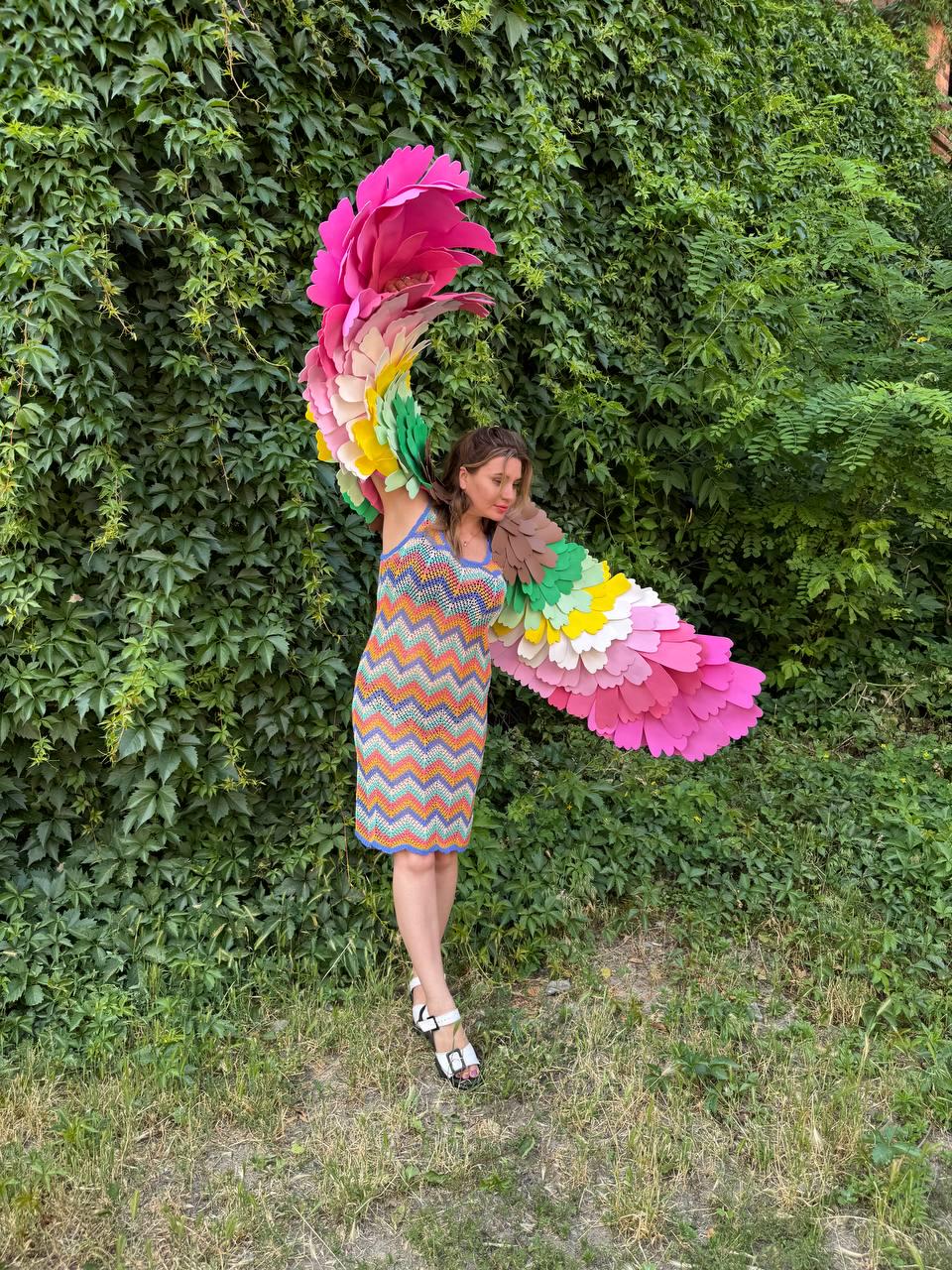 May include: A sleeveless chevron-patterned knit dress in orange, blue, yellow, and white, paired with white and black sandals and large, multi-colored fabric wings featuring layers of pink, yellow, green, and brown petal-shaped feathers.