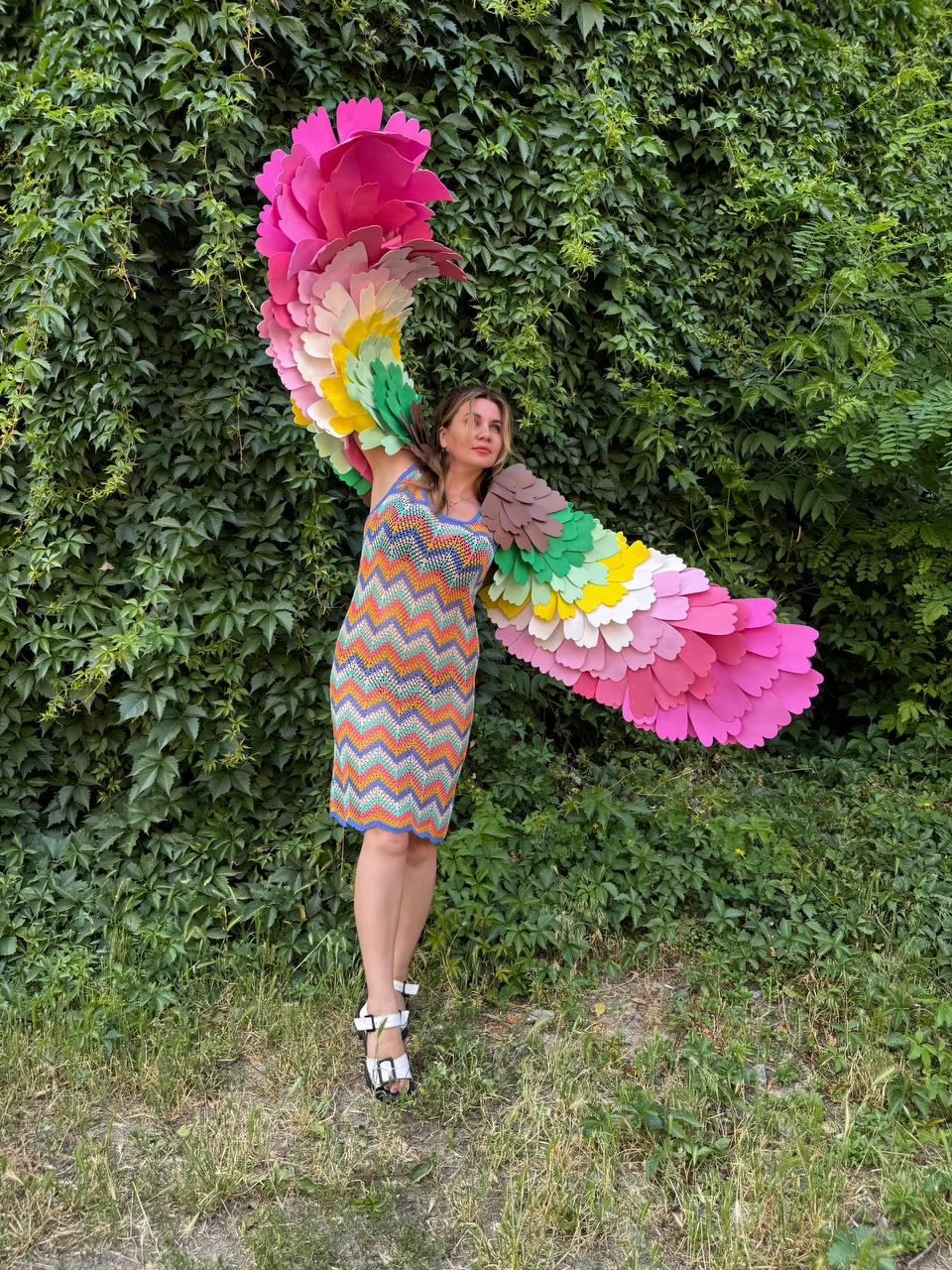 May include: A sleeveless chevron-patterned knit dress in orange, yellow, green, and blue, paired with white sandals. Large, expansive wings made of layered, scalloped felt feathers in a gradient of pink, yellow, green, and brown extend outward.
