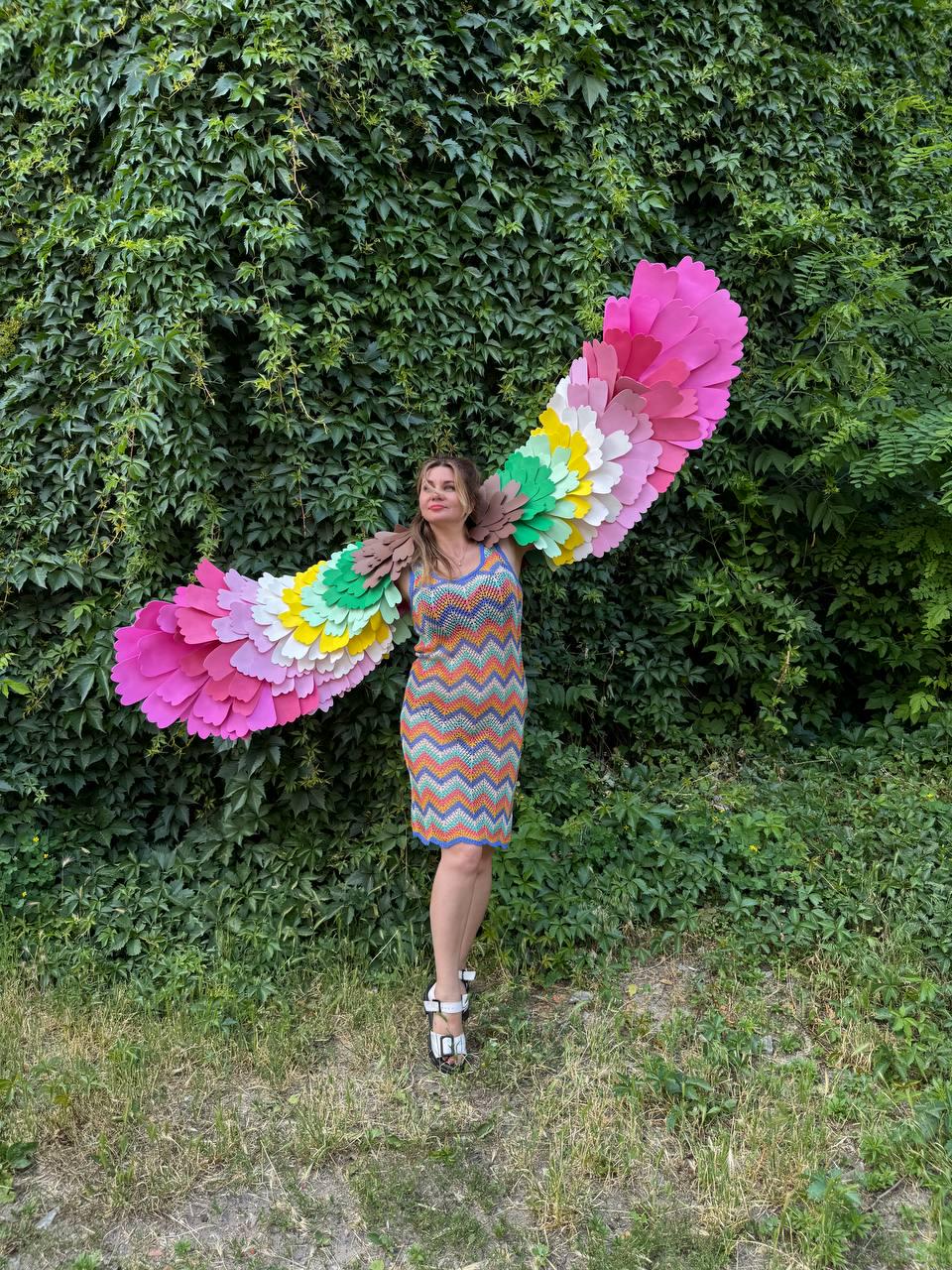 May include: A sleeveless chevron-patterned knit dress in orange, blue, and white, paired with large, expansive wings featuring layered, scalloped feathers in a vibrant gradient of pink, yellow, green, and brown, worn with white sandals.