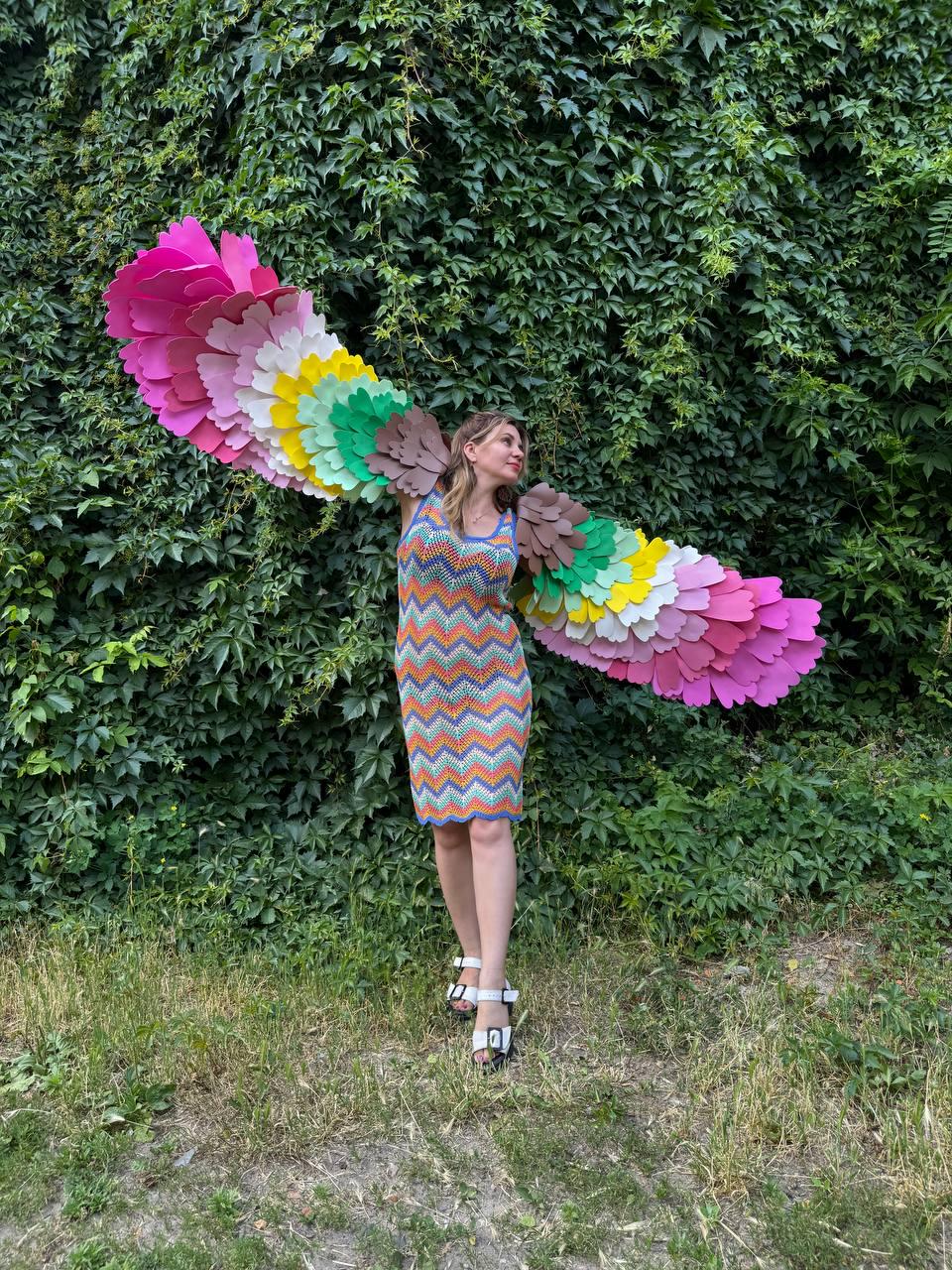 May include: A sleeveless knee-length dress featuring a multi-colored chevron pattern in orange, blue, and white. Attached to the back are large, expansive wings constructed from layered, petal-shaped fabric pieces in shades of pink, white, yellow, green, and brown.