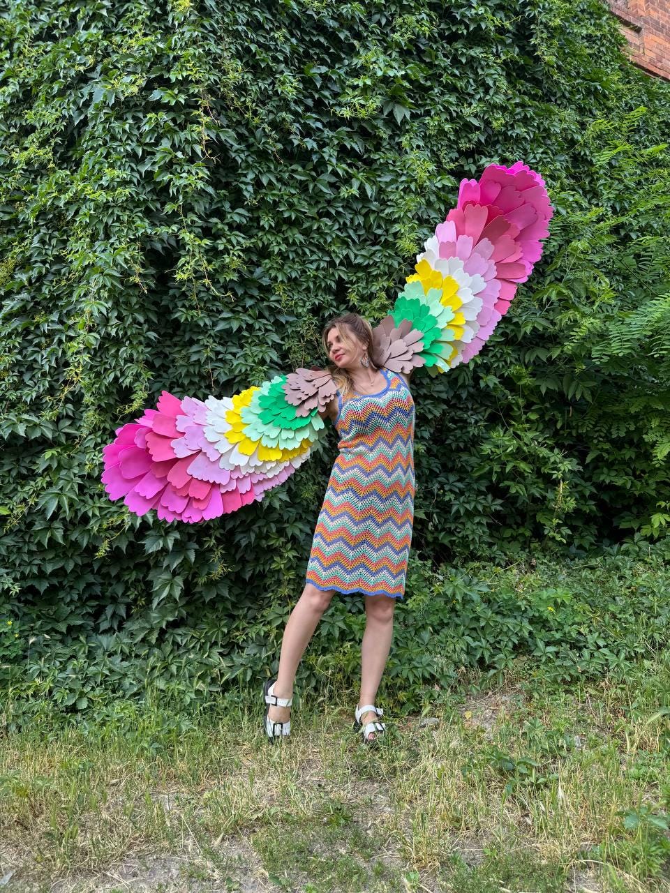 May include: A sleeveless knee-length dress featuring a multi-colored chevron pattern in orange, blue, and yellow. Attached to the back are large, expansive wings constructed from layered, scalloped felt feathers in shades of pink, yellow, green, and brown.