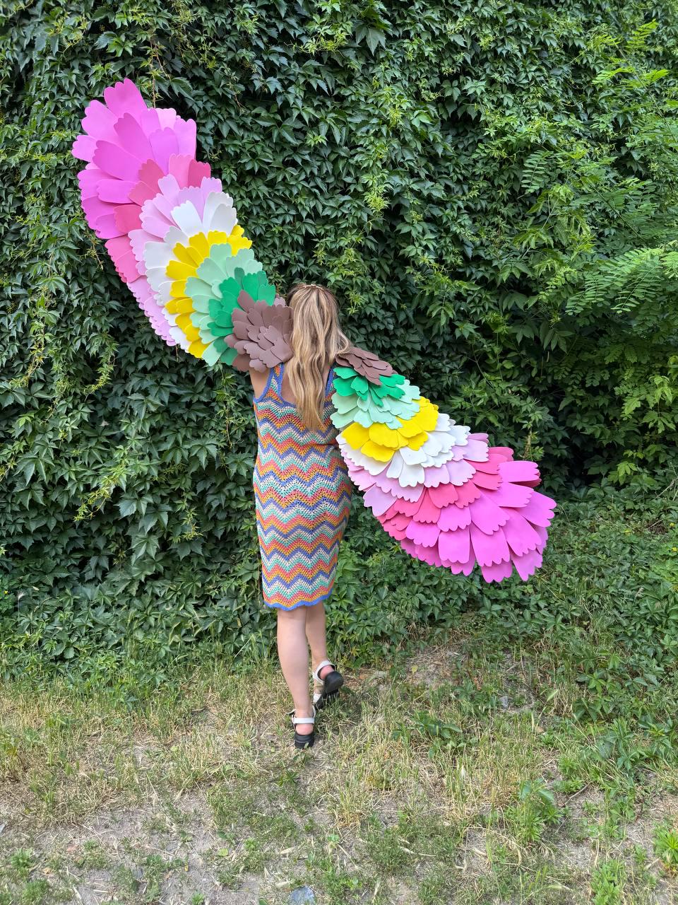May include: A sleeveless chevron-patterned knit dress in orange, blue, and yellow, paired with large, expansive wings constructed from layered, scalloped felt feathers in a vibrant gradient of pink, white, yellow, green, and brown hues.