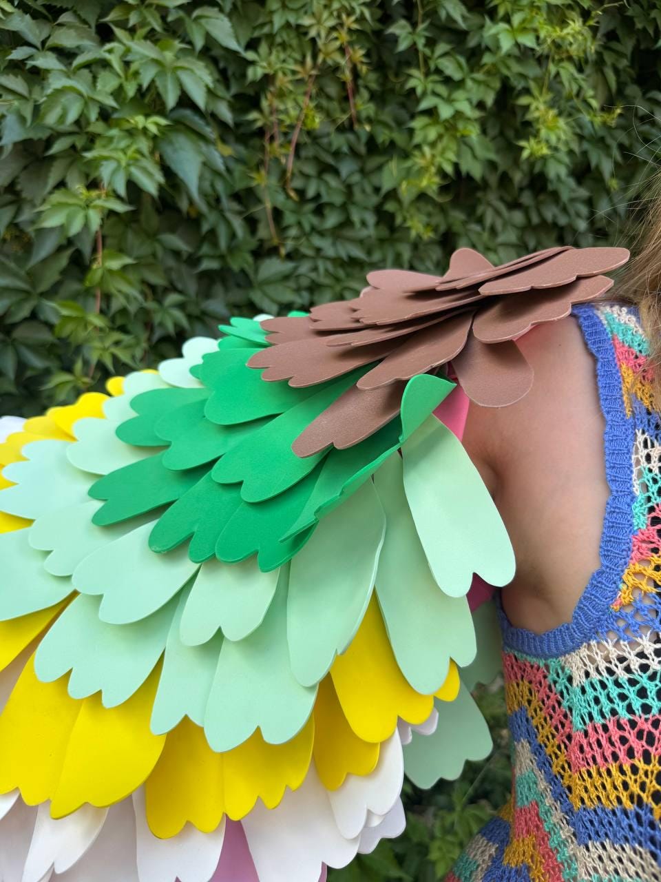 May include: A costume accessory featuring layered, scalloped foam petals in shades of brown, dark green, light green, yellow, and white, worn over a sleeveless, multi-colored crochet top with a blue trim and an open, net-like knit pattern.