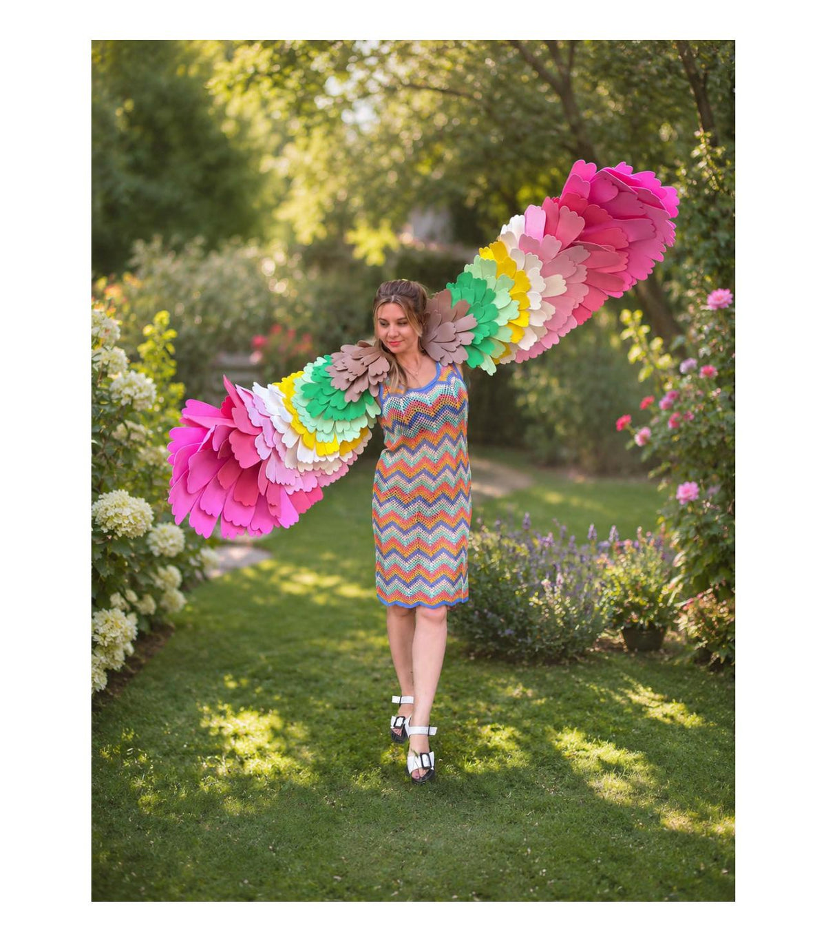 May include: A sleeveless chevron-patterned knit dress in orange, blue, and white, paired with large, expansive wings featuring layered, scalloped felt feathers in a vibrant rainbow gradient of pink, yellow, green, brown, and red hues.
