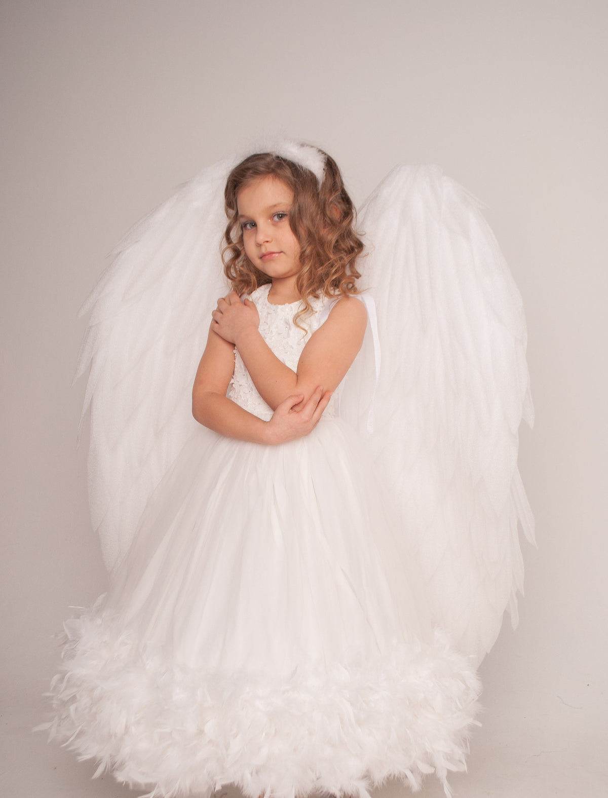 May include: A white dress featuring a sleeveless bodice with textured detailing and a full skirt trimmed with white feathers at the hem. Large, white, feathered angel wings are attached to the back, and a white feathered halo sits on the head.
