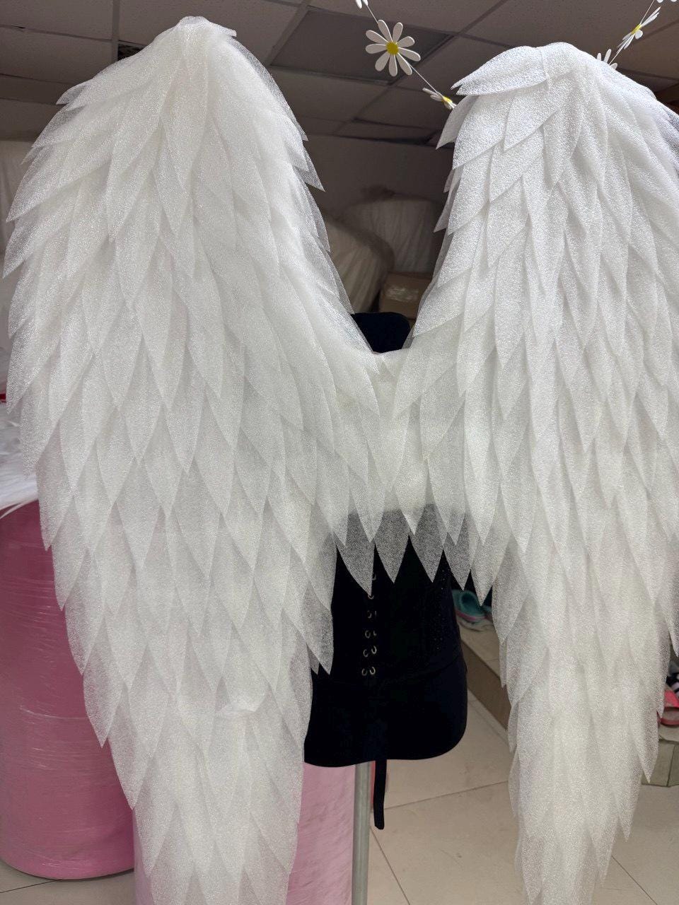 May include: A pair of large, white, glittery angel wings constructed from layered, leaf-shaped foam pieces. The wings are mounted on a black mannequin torso, displaying a textured, semi-translucent appearance with a shimmering finish across the surface.