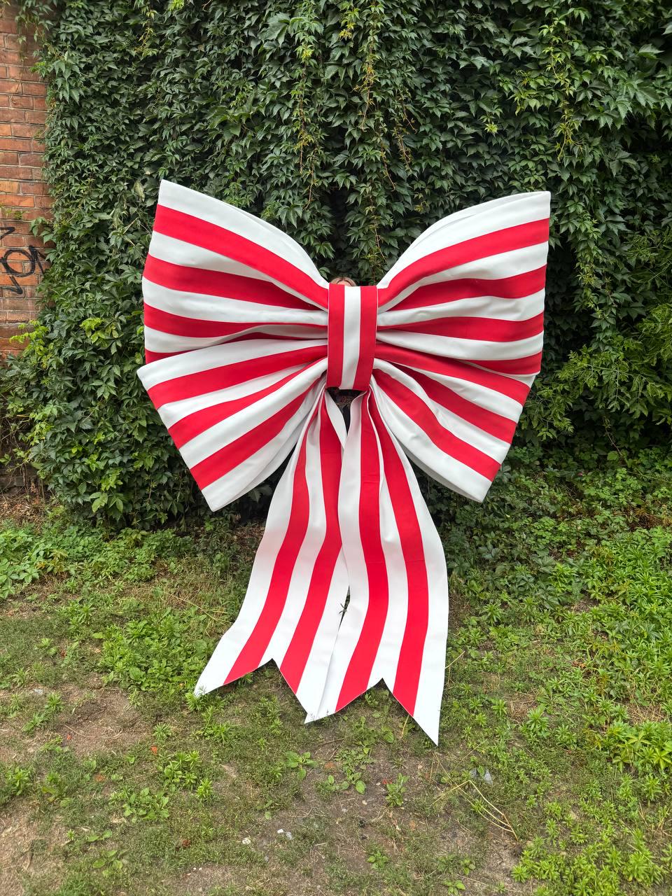 May include: A large decorative bow featuring a repeating pattern of bold red and white vertical stripes. The bow has two wide, symmetrical loops at the top and two long, flowing tails hanging down from the center knot, creating a classic shape.